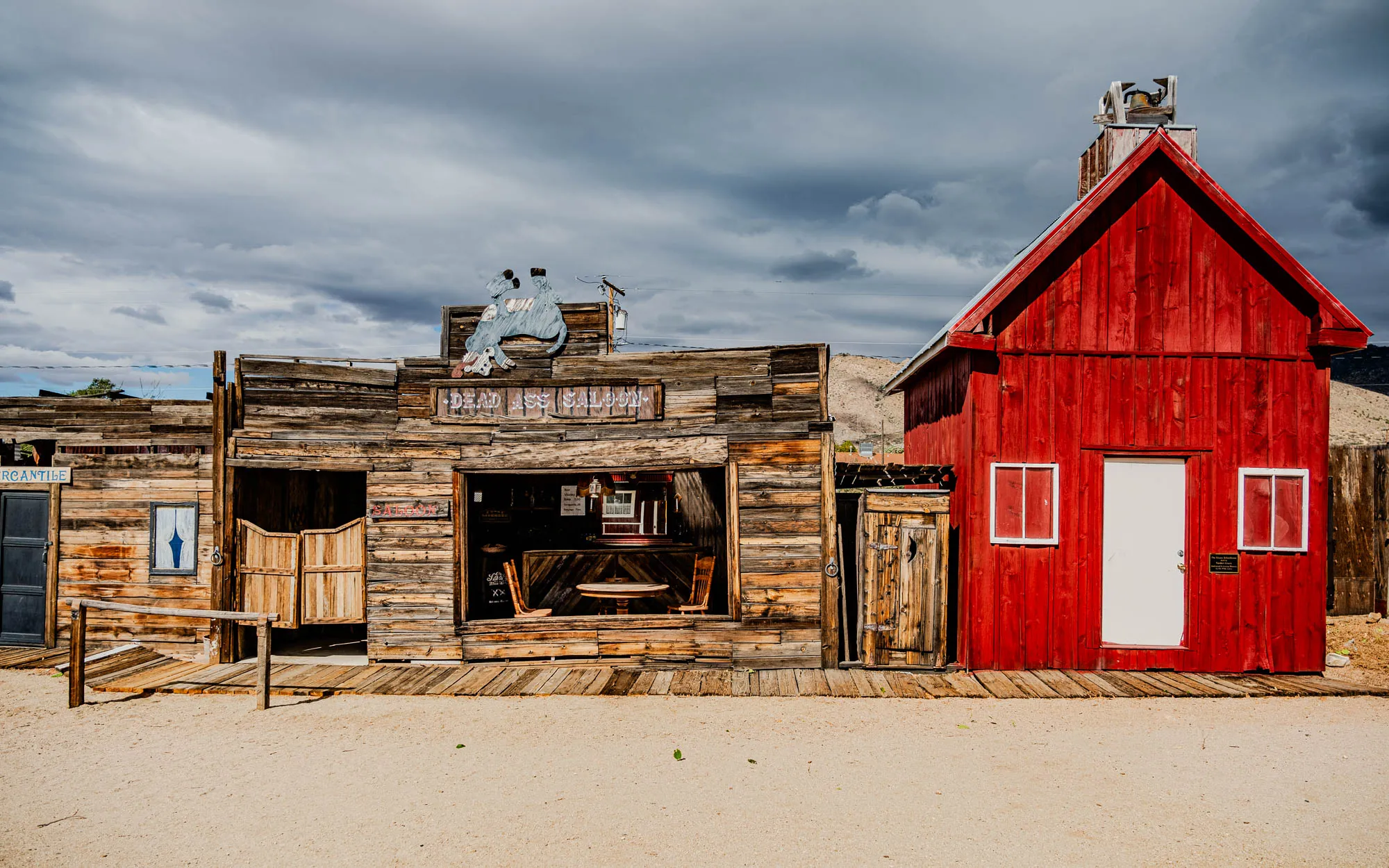 This image shows a western-style town with three wooden buildings. The building in the center is called "Dead Ass Saloon" and has a cutout of a bucking bronco above the entrance. The leftmost building is labeled "Mercantile" and has a dark blue door. The rightmost building is red, with a white door and two white windows with white frames. All the buildings are on a dirt road, with the sky above mostly overcast.