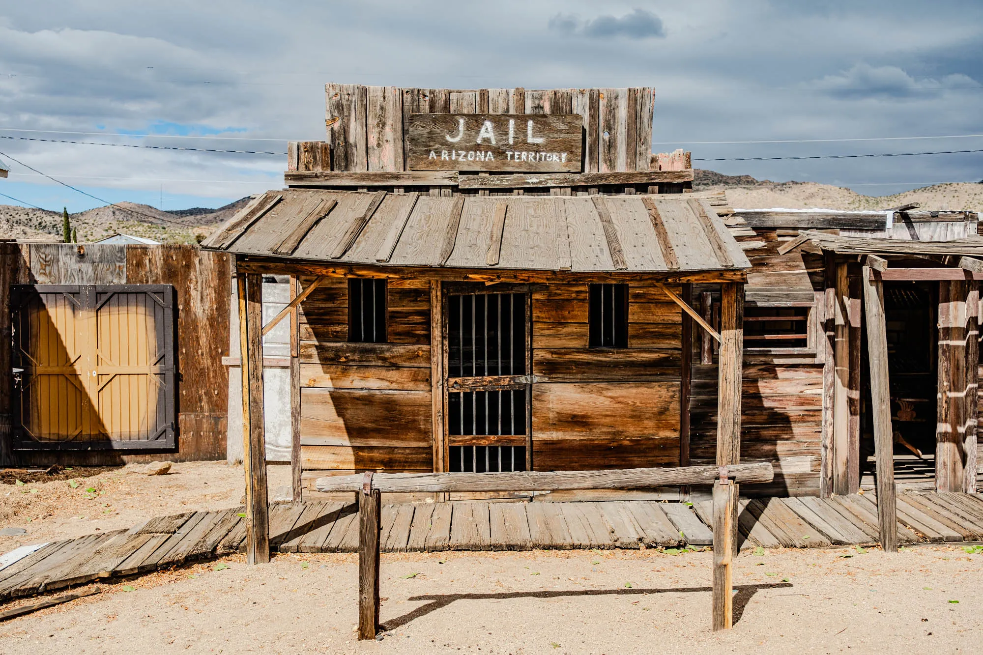 The image shows a wooden building with a sign above the entrance that reads "JAIL ARIZONA TERRITORY." The building is made of weathered wooden planks and has a wooden door with iron bars. The building has a roof made of wood planks and is surrounded by a wooden fence and a sandy area. The building has a rustic and aged appearance and gives the impression of being from the American West.  There is a wooden walkway leading to the front of the building. The sky is overcast with clouds, and there is a mountain range in the distance.  The image evokes a sense of history and frontier life.