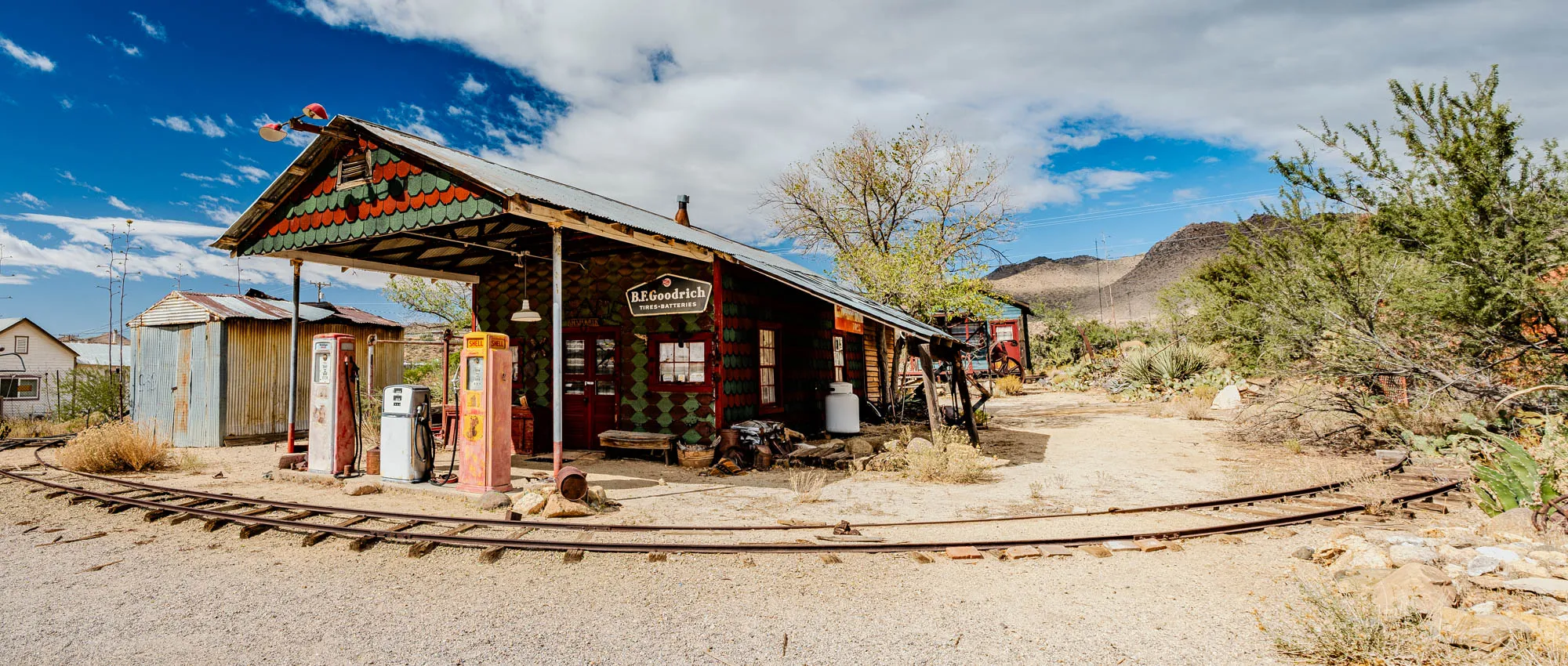 The image shows a weathered gas station in a desert landscape. The gas station has a green, red and brown striped roof and a sign that reads "B.F. Goodrich Tires-Batteries". There are two gas pumps and a rusty metal building to the left of the station. There are two sets of train tracks in the foreground. The image is taken under a blue sky with fluffy white clouds. There are dry, brown bushes and a large, dry tree in the background. There is a mountain in the distance.  