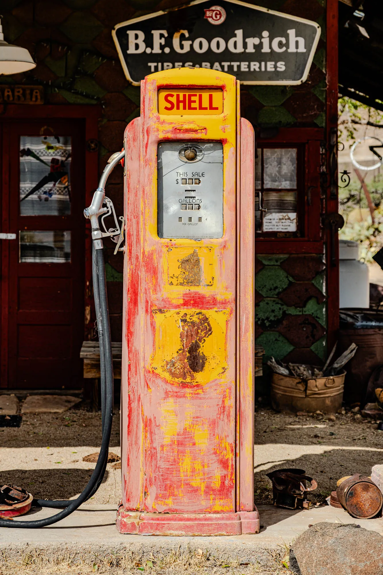 An old, red and yellow gas pump sits in front of a brick building. The pump is a bit faded and has some rust on it. It has a sign on the top that says "Shell". The price display is faded, but it looks like the price is $0.00. There is a sign above the pump that says "B.F. Goodrich Tires & Batteries". Behind the gas pump is a brown door. There are some old, rusty metal objects on the ground in front of the pump. The pump looks like it's from the 1950s or 1960s.