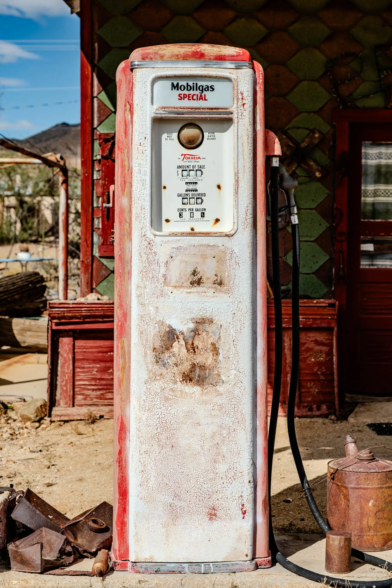 This is a photo of a vintage gas pump, a white metal rectangle with red accents and faded paint.  At the top of the pump it says "Mobilgas Special" and the words "Glass Must Be Full" and "Before and After Delivery". There are faded red numbers with "Amount of Sale" underneath.  "Gallons Delivered" is below that with blank spaces for numbers.  At the very bottom it reads "Cents per Gallon" with more blank spaces for numbers.  A black rubber hose runs from the side of the pump to the ground.  The pump is sitting in front of a building with red and green wooden siding with a door at the far right.  To the left of the pump is a red wooden box.   A rusted metal can sits in the lower right corner of the photo.