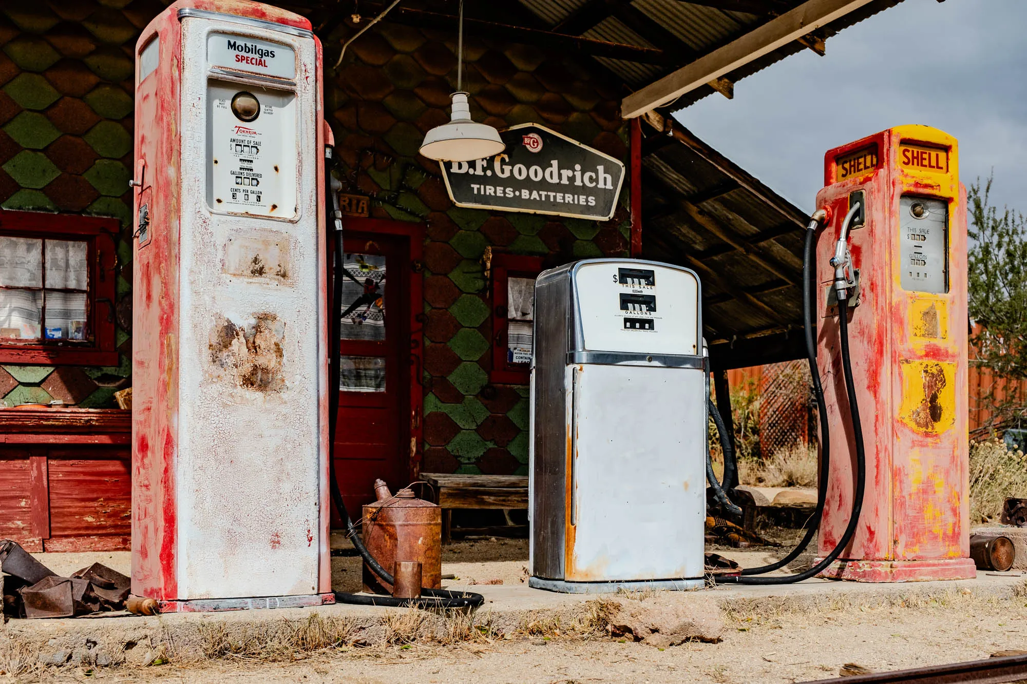 The image shows three vintage gas pumps, each painted in a combination of red, white, and yellow, standing in front of a weathered building.  The building has a sign above the door that reads "D.F. Goodrich TIRES BATTERIES." There is a light hanging from the roof of the building, and a metal watering can is sitting on the ground near the pumps. The image is a glimpse into a bygone era of gas stations. The first pump on the left has a white body, red trim, and a faded sign that reads "Mobilgas SPECIAL." The middle pump has a white body with a grey metal top and a sign that reads "This Sale." The third pump on the right is red with a yellow top and a sign that reads "Shell."  The pump handles are visible and they are made of black metal. The pumps are standing on a dirt and gravel ground.  There are a few small trees growing behind the pumps.  The image is taken from a slightly raised angle, giving the viewer a clear view of the pumps and building.