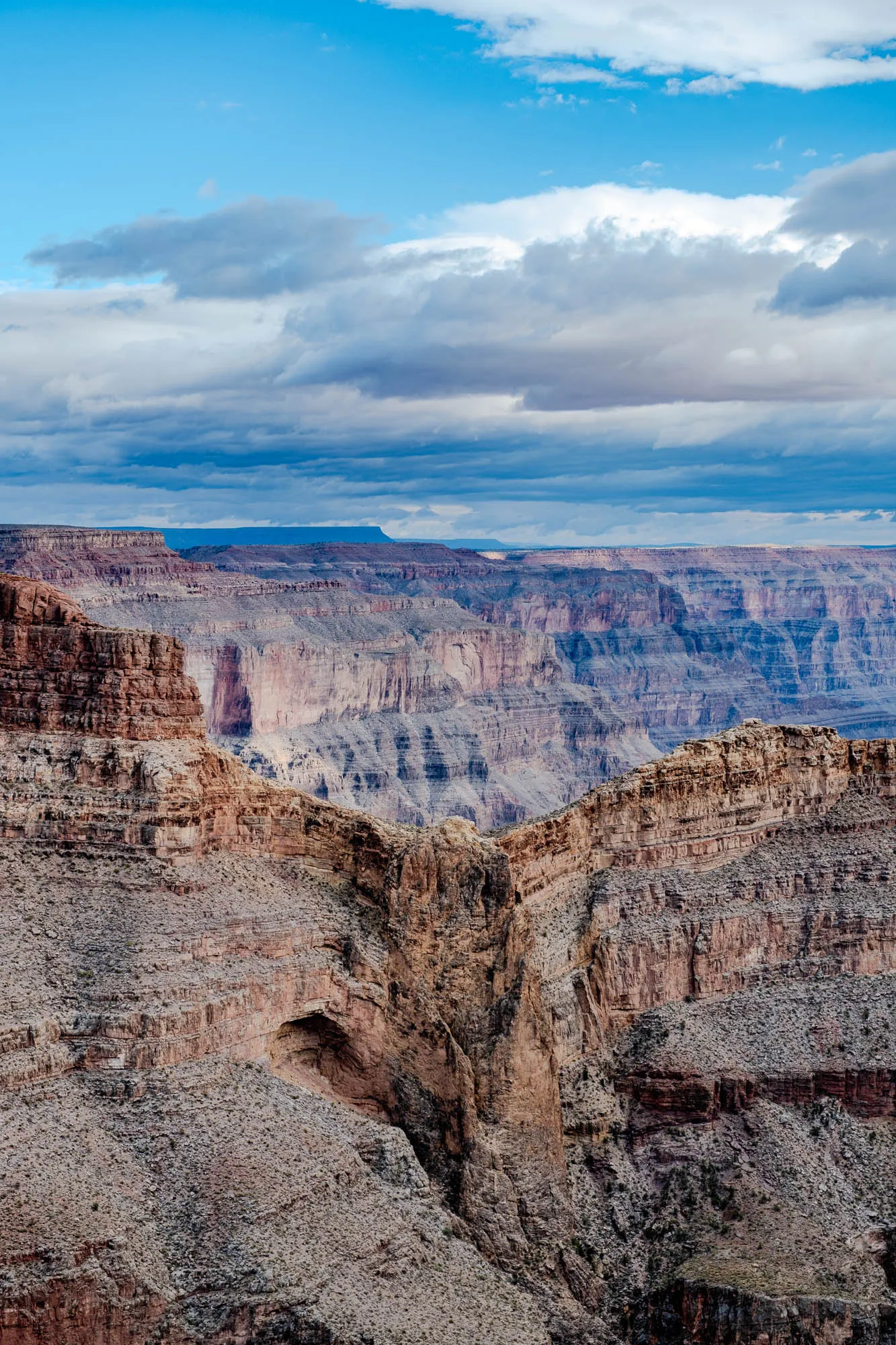 A panoramic view of a canyon. The top half of the image is a bright blue sky with puffy white clouds. The bottom half of the image is a steep, rocky canyon with layers of red, brown and grey rock. In the center of the image, the canyon dips down, creating a natural crevice. The canyon looks very large and deep, stretching out into the distance.  