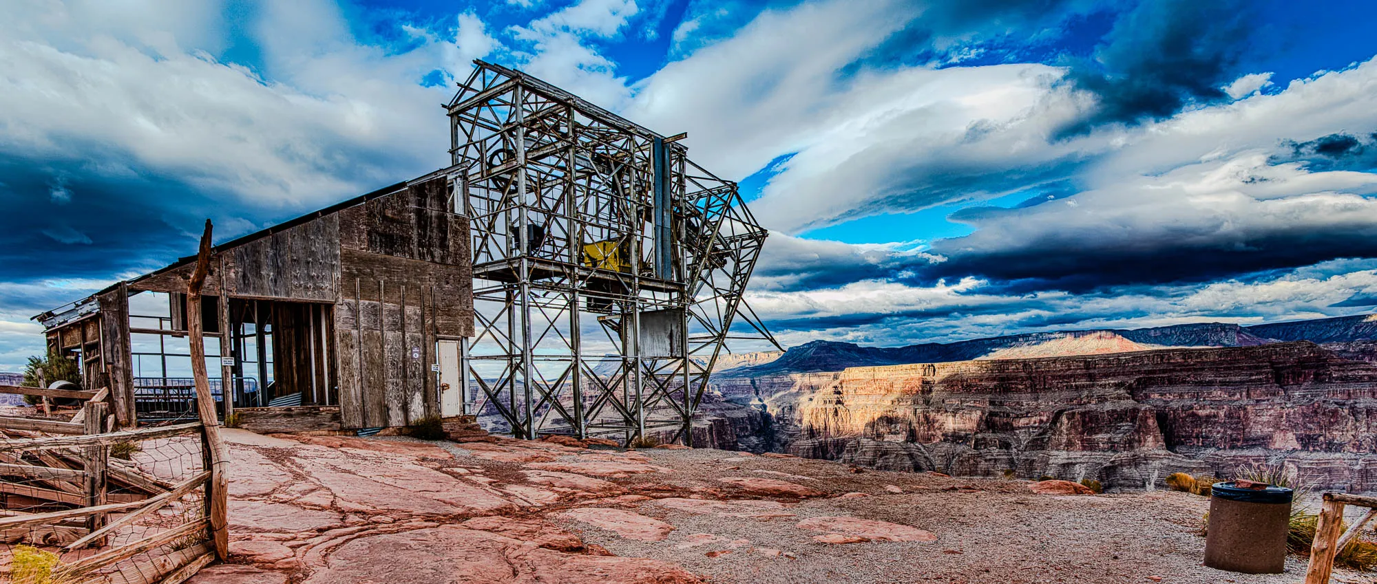 The image shows a large, rusty metal structure that looks like it could have once been a lift or a crane. There is a wooden building attached to the side of the structure. The building has a flat roof with a wooden plank sticking out from the side. The structure is perched on the edge of a cliff. There is a wooden fence on the left side of the image and a trash can on the right. The background is a deep canyon with layered rock faces. The sky is bright blue with large white clouds that are partially obscuring the sun. The image is taken from a low angle, looking up at the structure. The colors are muted and the overall feeling is one of solitude and decay.