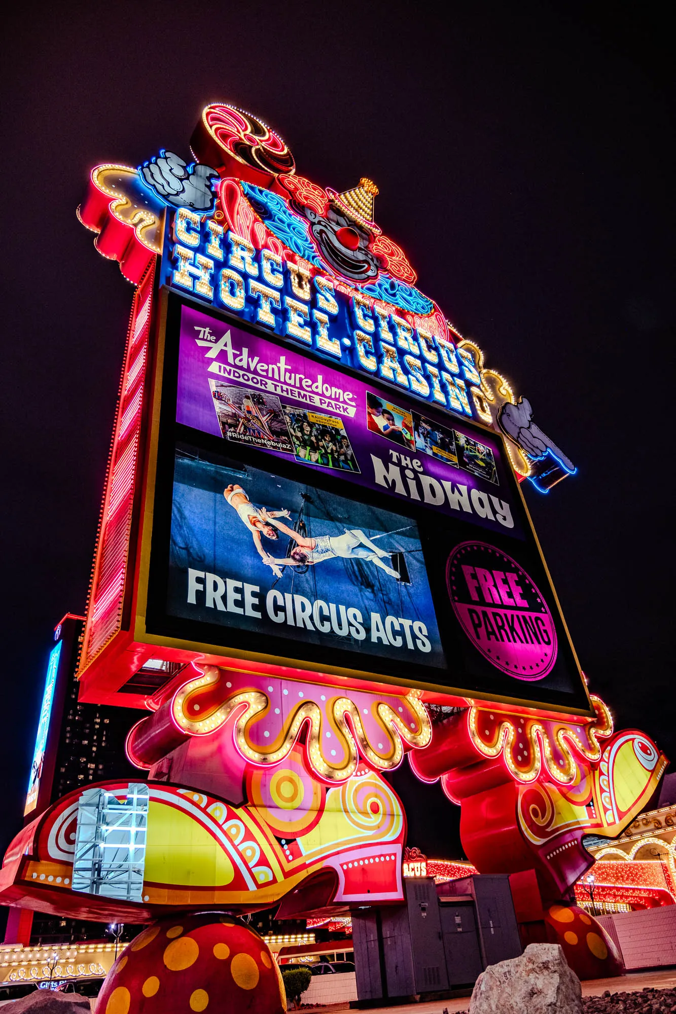 The image is a night shot of a giant, colorful neon sign for a hotel and casino. The sign is designed in the shape of a clown with a giant red nose, large, bright, white eyes, and a big smile. The sign reads "Circus Circus Hotel Casino" in glowing, neon letters. The clown's arms are raised above its head. The clown is standing on oversized, cartoon shoes with yellow and pink polka dots and swirls.  The sign also has a smaller, rectangular advertising space with three framed images and text that reads "The Adventuredome Indoor Theme Park," "The Midway," "Free Circus Acts," and "Free Parking." The sign is illuminated in a variety of bright colors and is surrounded by darkness, highlighting its vibrancy. The sign is set against a background of other buildings.  You can see the top of a building with a "Gifts" sign on its side.