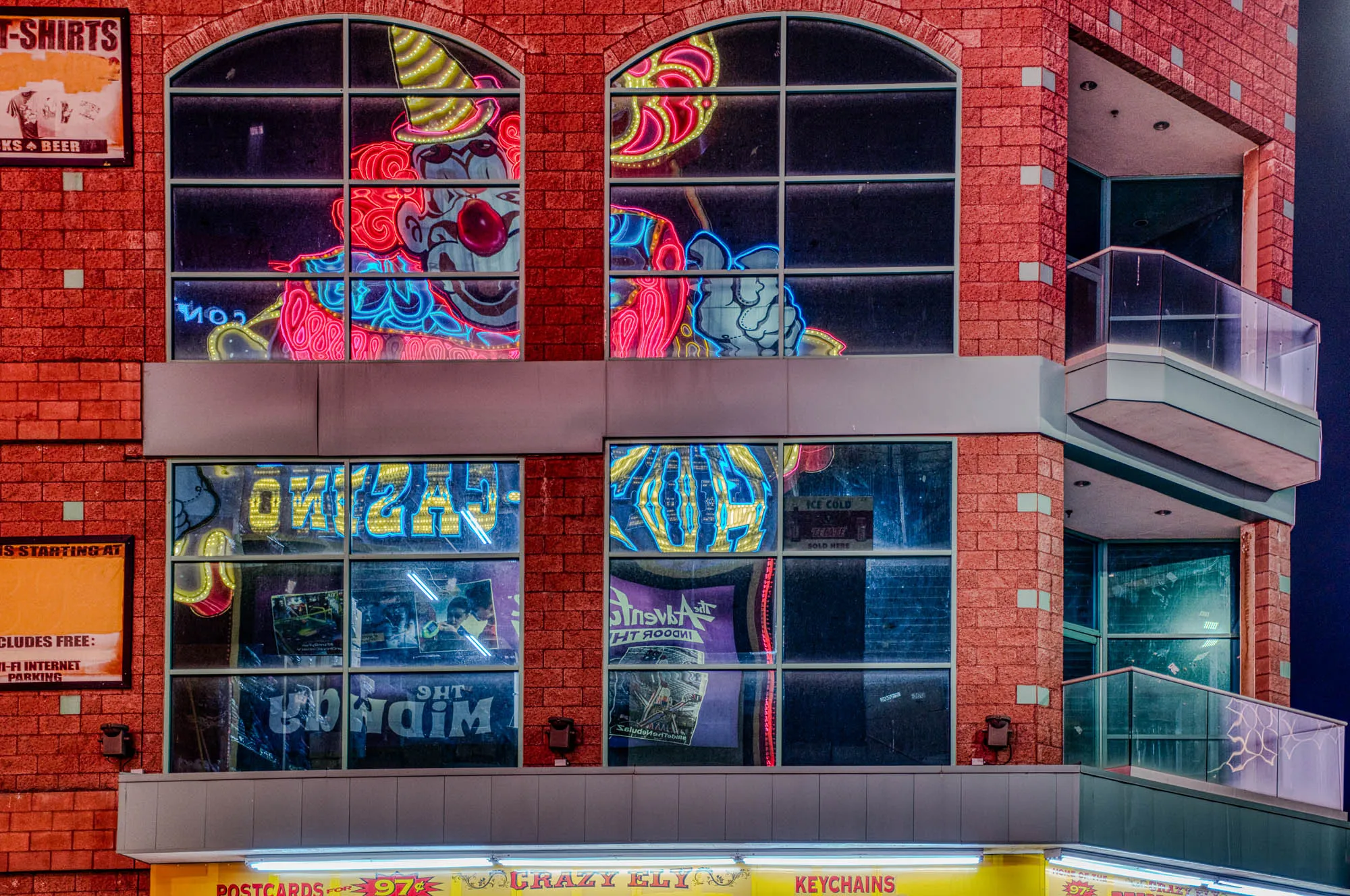 The image is of a brick building with a large window at the top. In the window, there is a neon sign of a clown, with a red nose, a red smile, and a blue hat with a yellow star on it. The clown is wearing a red and white outfit. The windows are divided into multiple panes. There are other signs in the windows below, including a sign that says "Crazy Eli" and one that says "Postcards." There is a small balcony on the side of the building, which is also made of brick. There are two signs at the bottom of the image that say "Postcards for 97¢" and "Keychains." The background is a dark blue.  There is a yellow sign with the word "T-shirts" on the left side of the image. Below the sign, in small font, it says "KS Beer".  To the right of the "T-shirts" sign is a small, orange sign that says "CLUDES FREE" and "WI-FI INTERNET" and "PARKING". The image is a view of a tourist area, possibly in Las Vegas, Nevada.