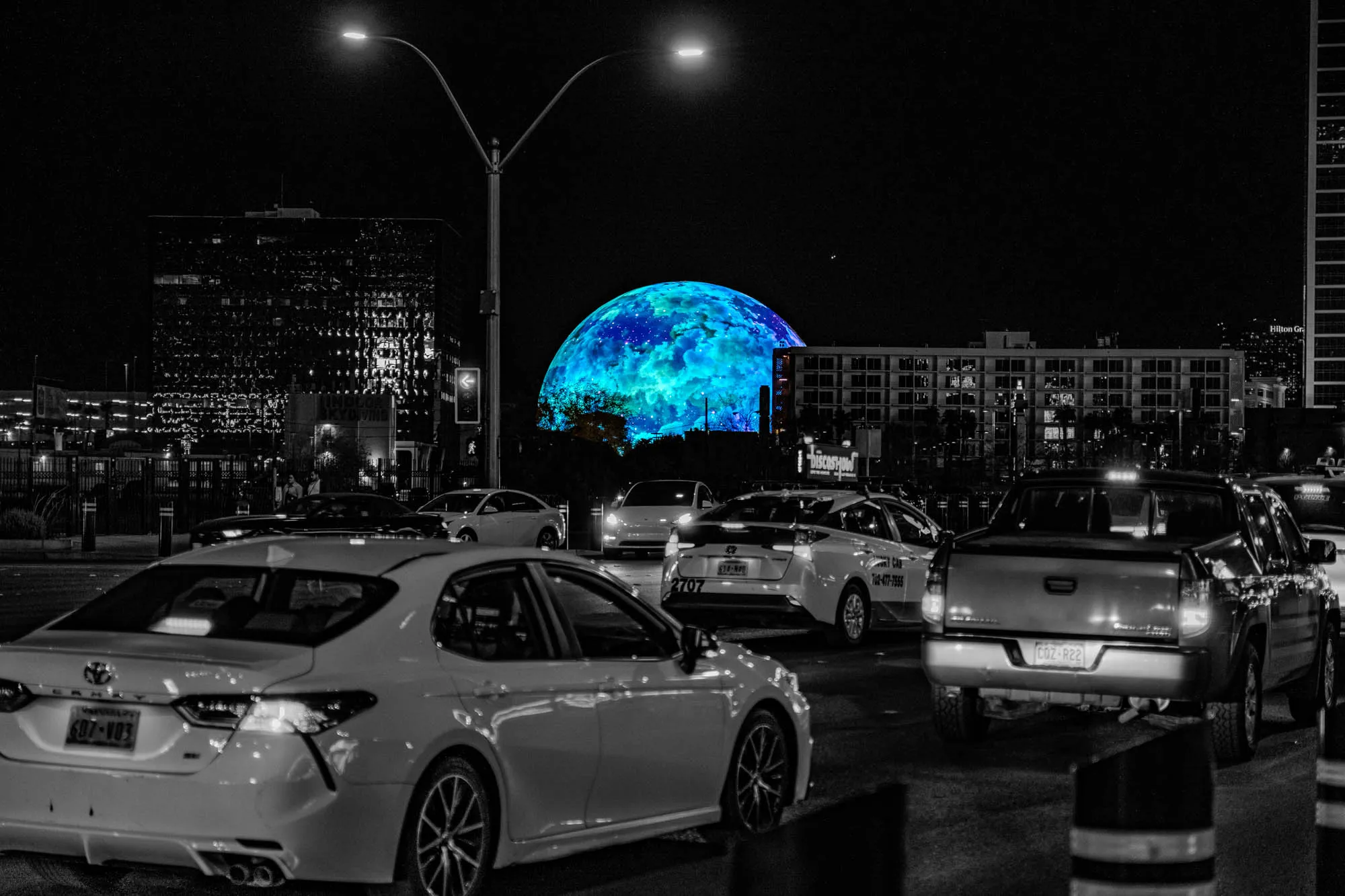 The image is a black and white photo of a city street at night. In the center of the image, there is a large, blue and green, sphere shaped structure. It is lit up and appears to be a digital projection. It's surrounded by several buildings with windows.