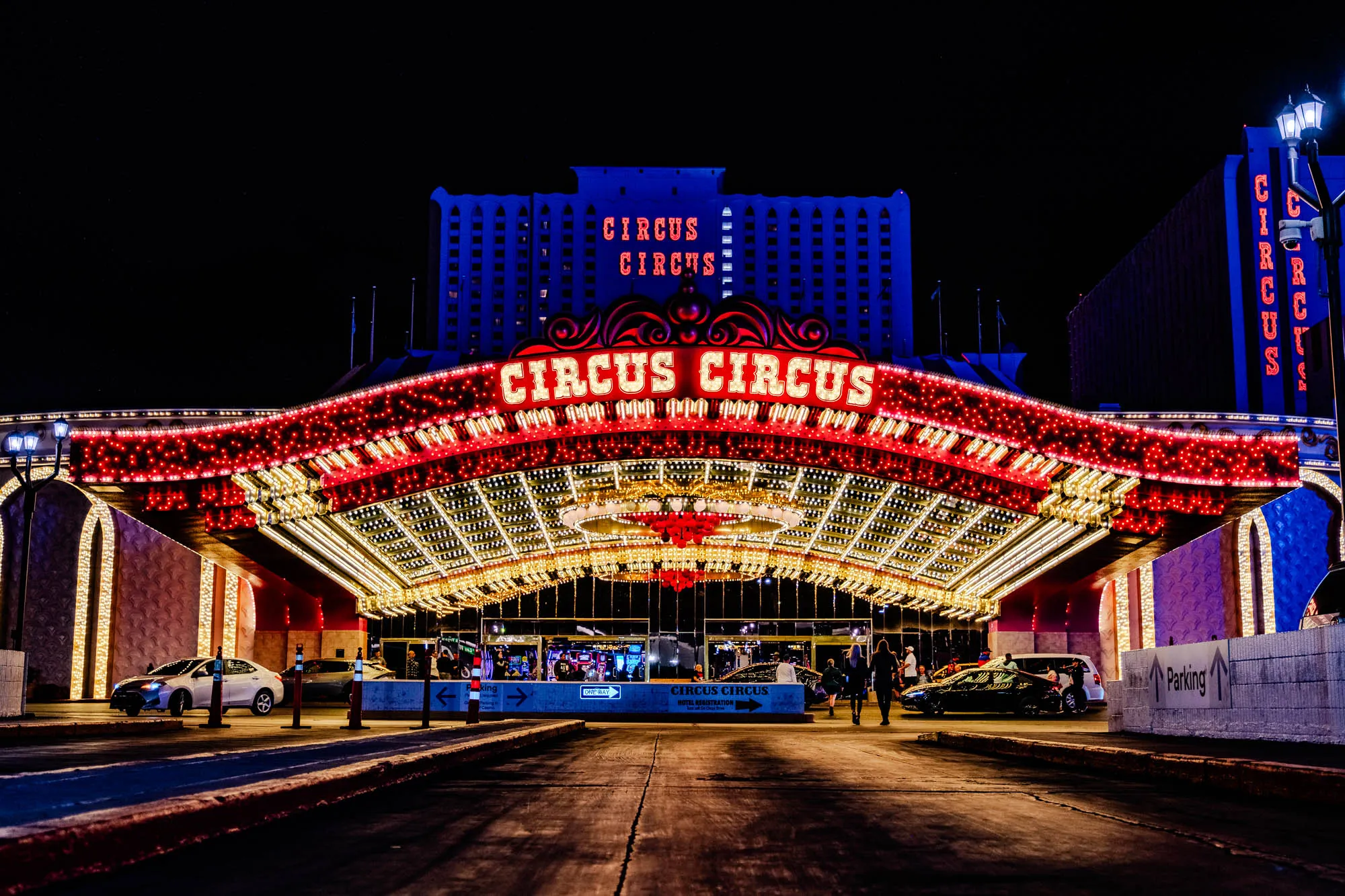 The image shows a nighttime view of the Circus Circus hotel and casino in Las Vegas. The entrance to the casino is a large, arched structure with a neon sign that reads "Circus Circus" in red letters. The sign is adorned with festive lights, creating a bright and inviting atmosphere. 

The hotel itself is a tall, multi-story building with a white facade and rows of windows. The building also has a large neon sign that reads "Circus Circus" in red letters. 

There are people walking around the casino entrance, and several cars are parked nearby. The ground is paved, and there are some concrete barriers and directional signs. 

The scene is vibrant and bustling, and it creates a sense of excitement and entertainment. The image is typical of the Las Vegas Strip and its iconic atmosphere. 
