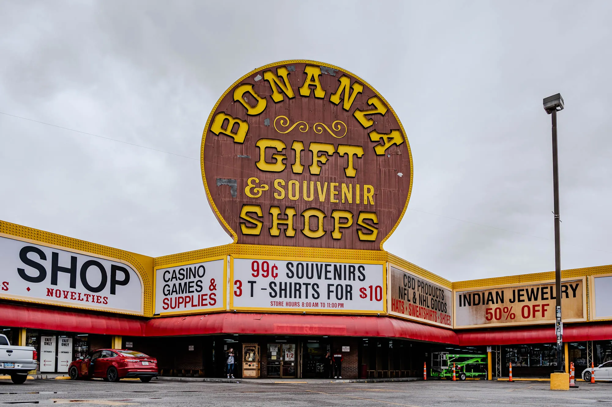 The image shows a storefront with a large, round sign that reads "Bonanza Gift & Souvenir Shops". The sign is yellow and brown with ornate yellow lettering. The storefront is divided into several sections, each with its own sign. The leftmost section is a "Shop Novelties" store. The middle section is a "Casino Games & Supplies" store, and the rightmost section is a store selling "Indian Jewelry, 50% Off".  The storefront has a large, red awning and is fronted by a parking lot with a few cars. There is a tall black pole in the background with a streetlight at the top. In the center of the image there is a sign that reads "99¢ Souvenirs, 3 T-shirts for $10", with the words "Store Hours 8:00 am to 11:00 pm" below.  There is also another sign advertising "CBD Products, NFL & NHL Goods, Hats, Sweatshirts, Shoes". There are a few people walking around in the parking lot. The sky is a cloudy gray.