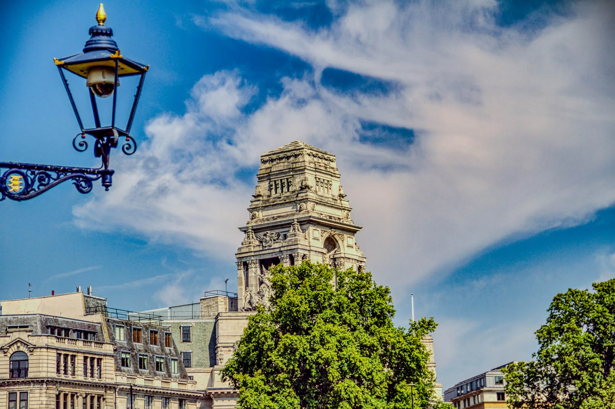 The image shows a tall, ornate building with a tower. The tower has decorative details and is topped with a dome. The building is made of stone and has many windows. There is a large tree to the right of the tower. The image is taken from a low angle, with a street lamp in the foreground. The lamp is black and gold and has a ornate design. Inside the lamp is a surveillance camera. The sky is blue with white clouds. The scene is bright and sunny. There is also a smaller building behind the tall building. The smaller building is a flat structure, with a grey roof.