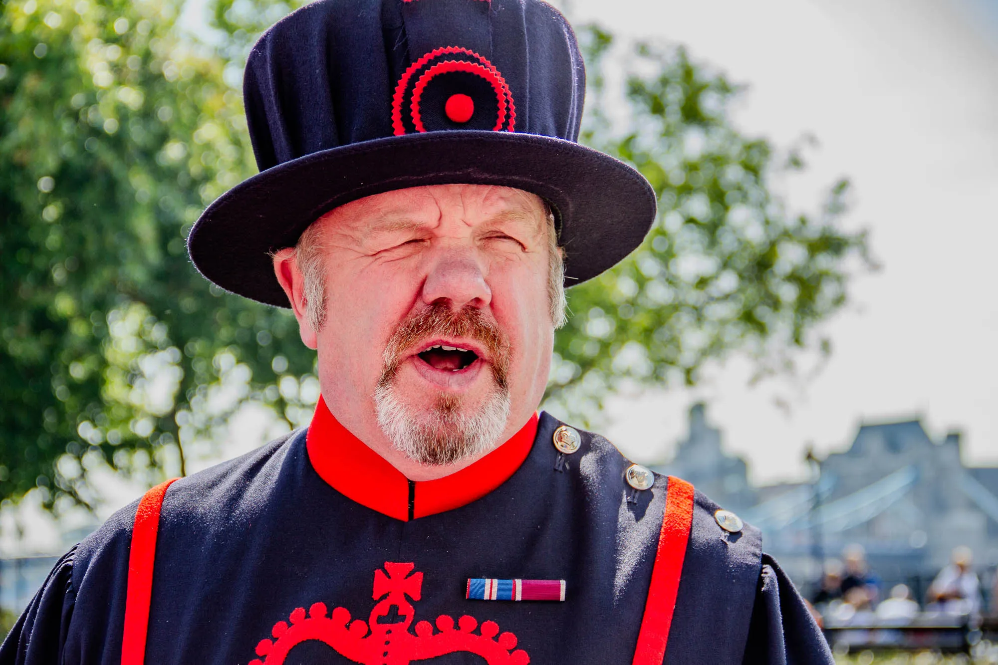 The image shows a man wearing a black and red uniform and a large black hat with a red circle emblem. He has a white beard and mustache. His mouth is open as if he is speaking. There is a red ribbon with a silver badge and another small badge on his uniform. The man is in front of a blurry background that appears to be a green tree, possibly a park, and an out-of-focus tower bridge.