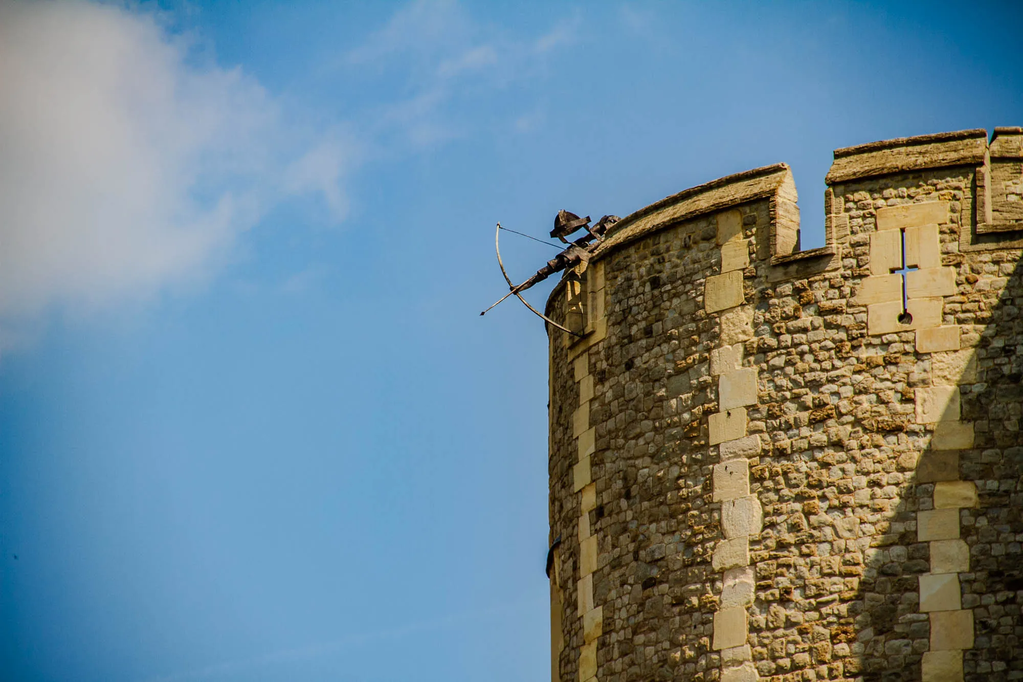 The image shows a section of a tall stone tower against a bright blue sky with a few fluffy white clouds. The tower is made of rough, brown stones, and there is a large, white, rectangular window on the side. Atop the tower, there's a metal sculpture of a person holding a bow and arrow. The bow is pointed towards the ground. The sculpture is partially obscured by a wall, so only the head, bow, and arrow are visible. The wall casts a dark shadow on the tower.