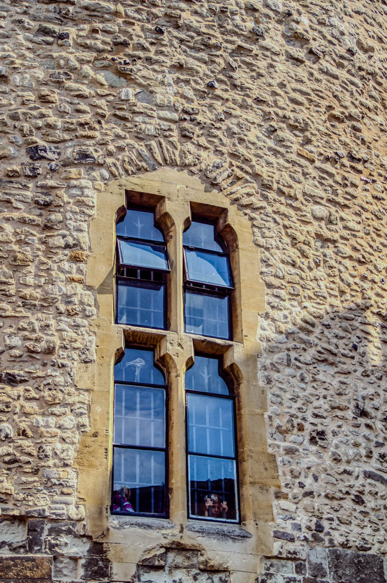 This image shows a window in a stone wall. The wall is made of rough, irregular stones. The window has four panes, all with black frames. Two of the panes are open, with the bottom portion hinged to open outward. Through the window, you can see two people.  The person on the left is wearing a purple jacket, and the person on the right is holding their hands to their face.  The window appears to be on the upper level of a building, as there is no other structure in the photo, only the stone wall, which is likely part of a tower or castle.  The image is taken on a sunny day.