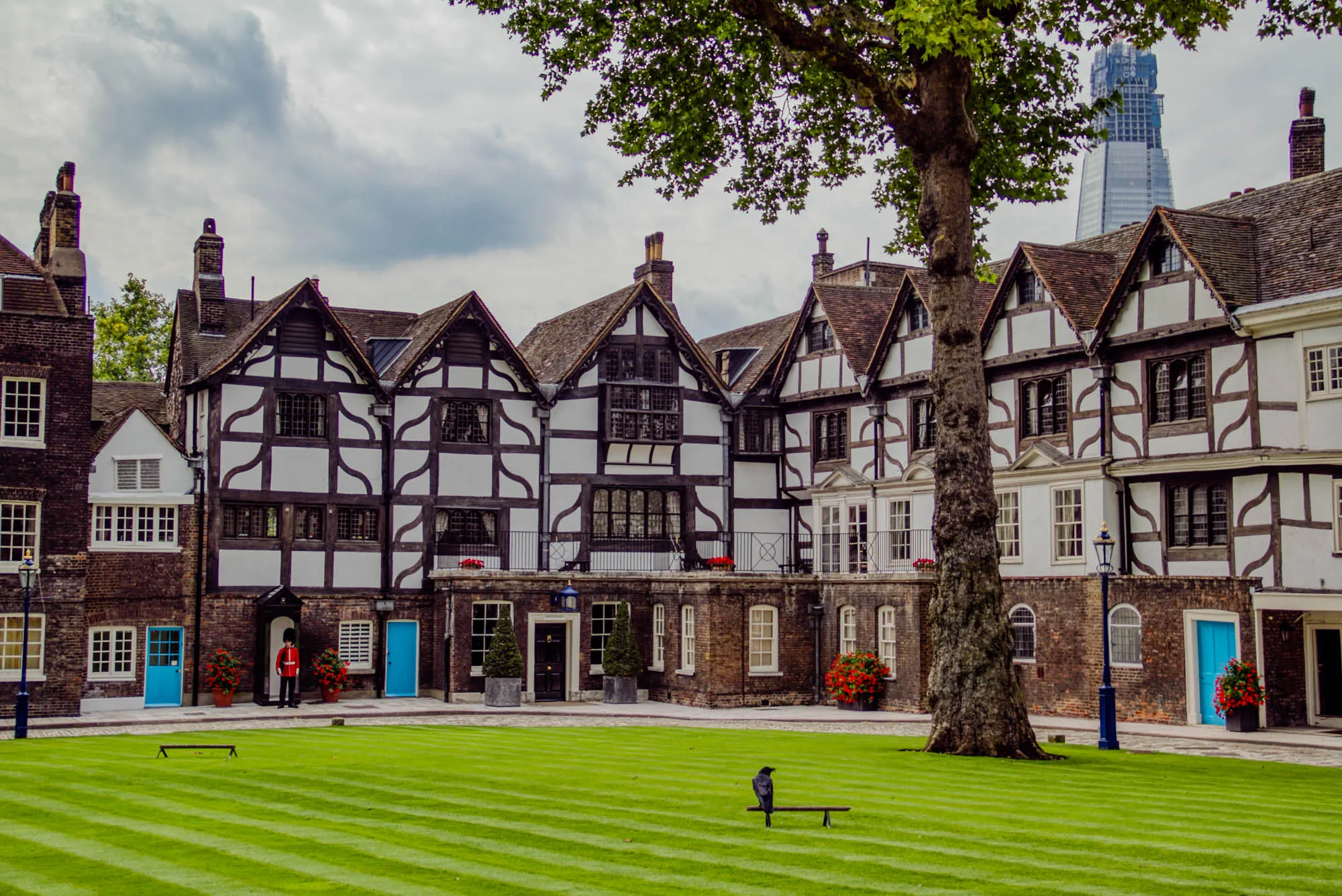 The image shows a courtyard with a large, old, brick building with a white half-timbered facade. The building has many windows and a roof that is visible only partially. To the left of the building, in the background, there is a second, brick building with white windows. Both buildings are flanked by green trees. In the foreground there is a large, green, manicured lawn with a black crow standing on a dark, wooden bench. The lawn is striped by rows of evenly cut grass.  To the right of the lawn there is a large tree that partially obscures a modern, glass skyscraper in the background. In the foreground, there is a person in red and black uniform standing in front of a blue door. There is a black streetlamp to the left of the image and another one to the right of the image.