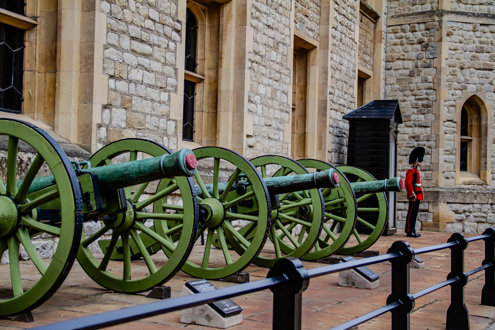 The image shows five large, green, wooden cannons lined up in a row along the wall of a castle. The cannons have spokes like a wheel and have black metal bands wrapped around them. The cannons are aligned from left to right with the wheels slightly overlapping. Behind the cannons, a stone wall has a window and a black door. In the right side of the image, a person wearing a red uniform and a black fur hat stands facing the cannons. The person is a member of the British Royal Guard. The ground in front of the cannons is made of brown bricks and has a black metal railing.