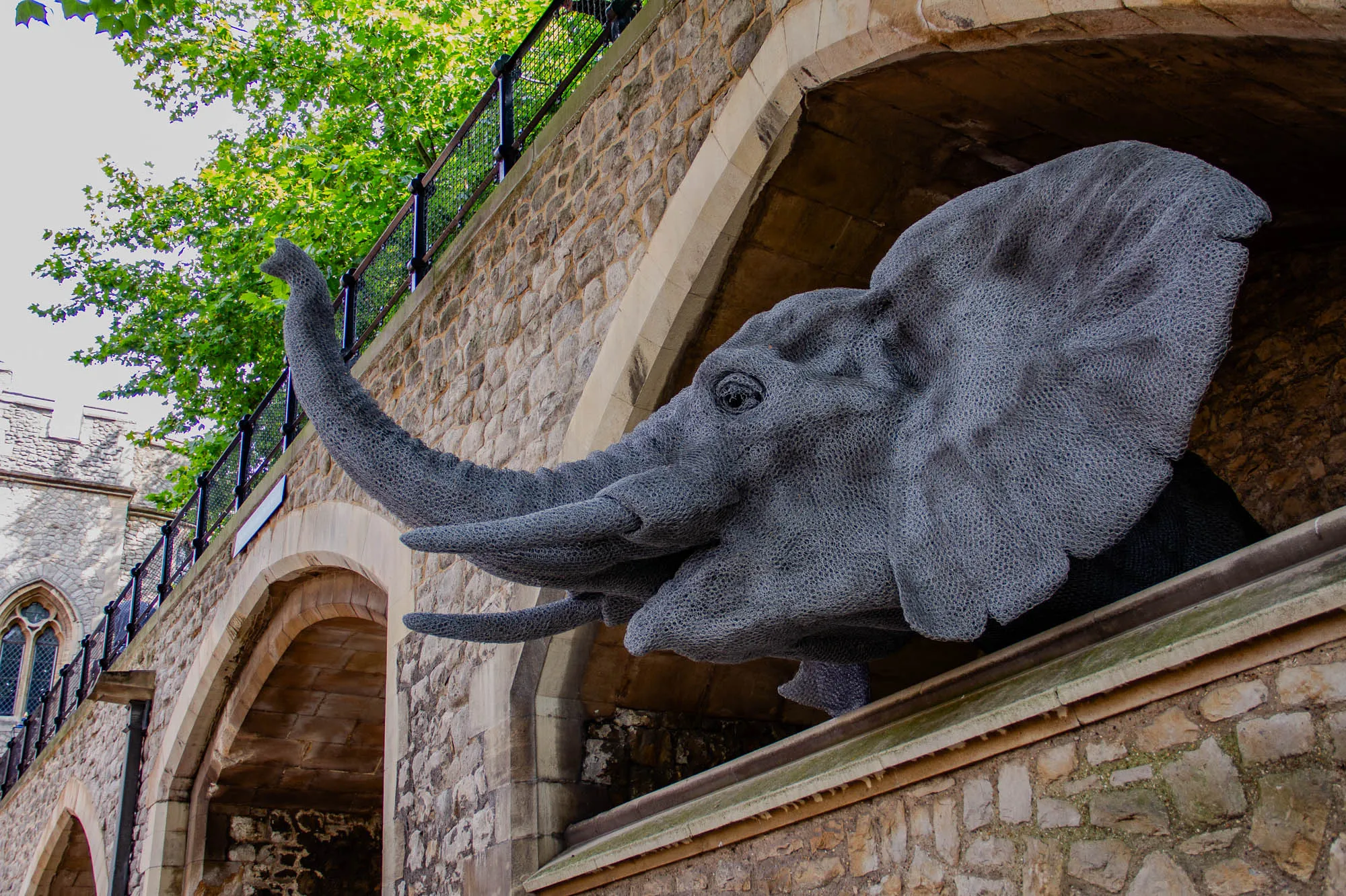 A close-up of a large metal sculpture of an elephant's head. The elephant is made of wire mesh, and its head is sticking out from an archway. The elephant's trunk is raised in the air, and its eyes are closed. The sculpture is made of a grey metal, and it is set against a backdrop of a stone wall. There are trees and a stone wall and metal railing in the background. 