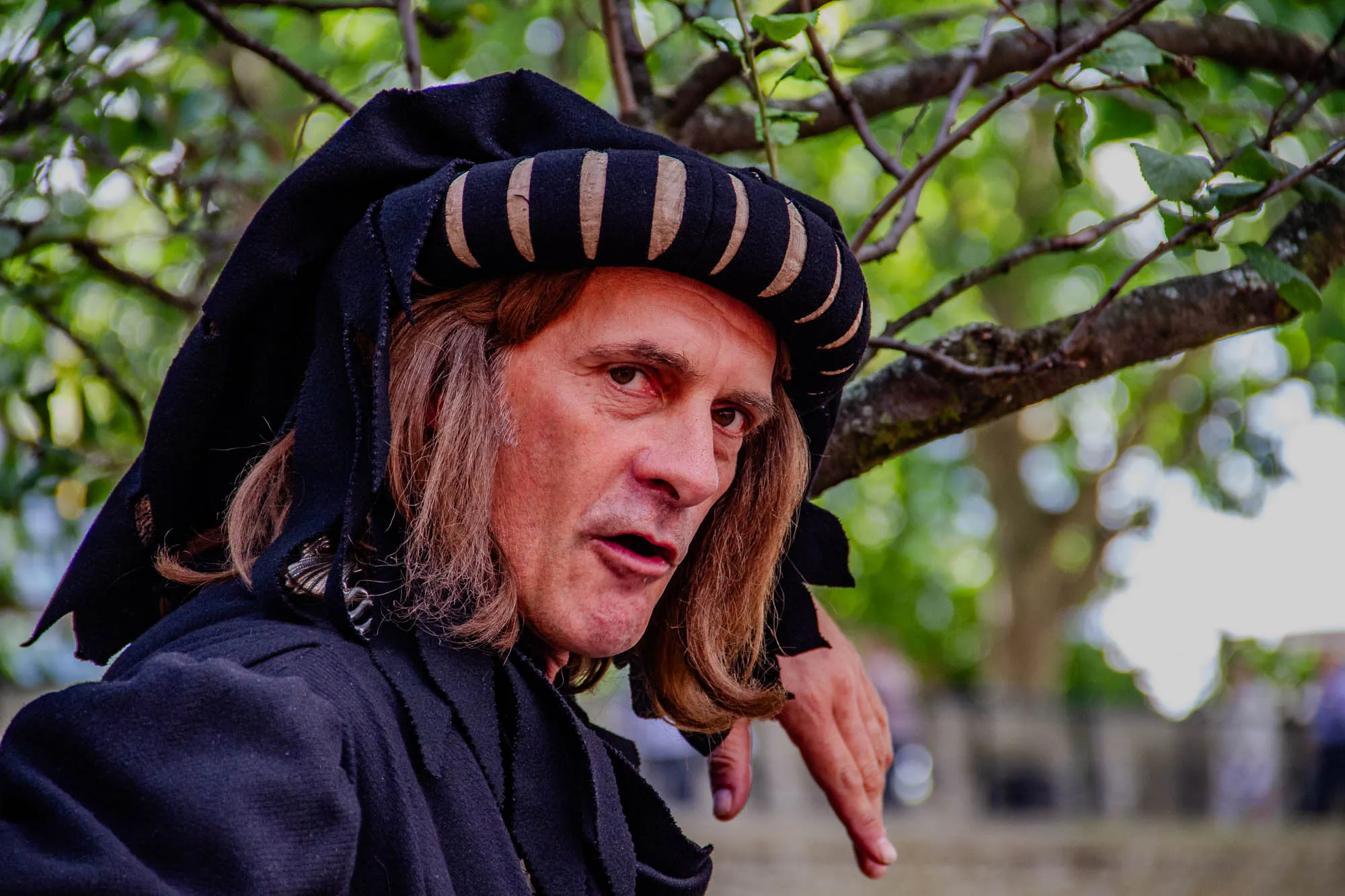 A man with long brown hair is wearing a black, hooded,  medieval-style tunic with a brown and black striped band on the hood.  His eyes are brown, his skin is fair,  and he has a light beard and mustache. He is looking toward the left of the frame, his mouth is slightly open, and his right hand is resting on a branch. The man is in focus, and a blurry background suggests a wooded area. 