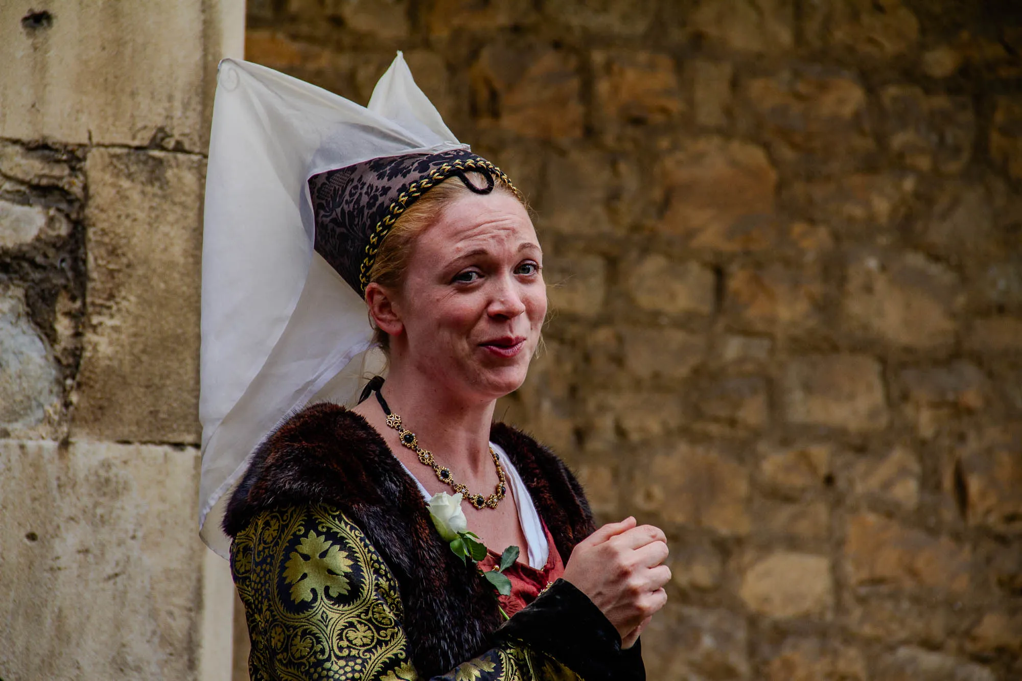 The image shows a woman wearing a white, hooded gown with a brown fur trim, and a black and gold patterned dress underneath. She is wearing a gold chain with dark gemstones around her neck. She is smiling and has her hands clasped together in front of her. She is standing in front of a rough stone wall. The woman's hair is red and she is wearing a black and gold patterned headdress. She is looking directly at the viewer. The image is likely taken during a historical reenactment.  
