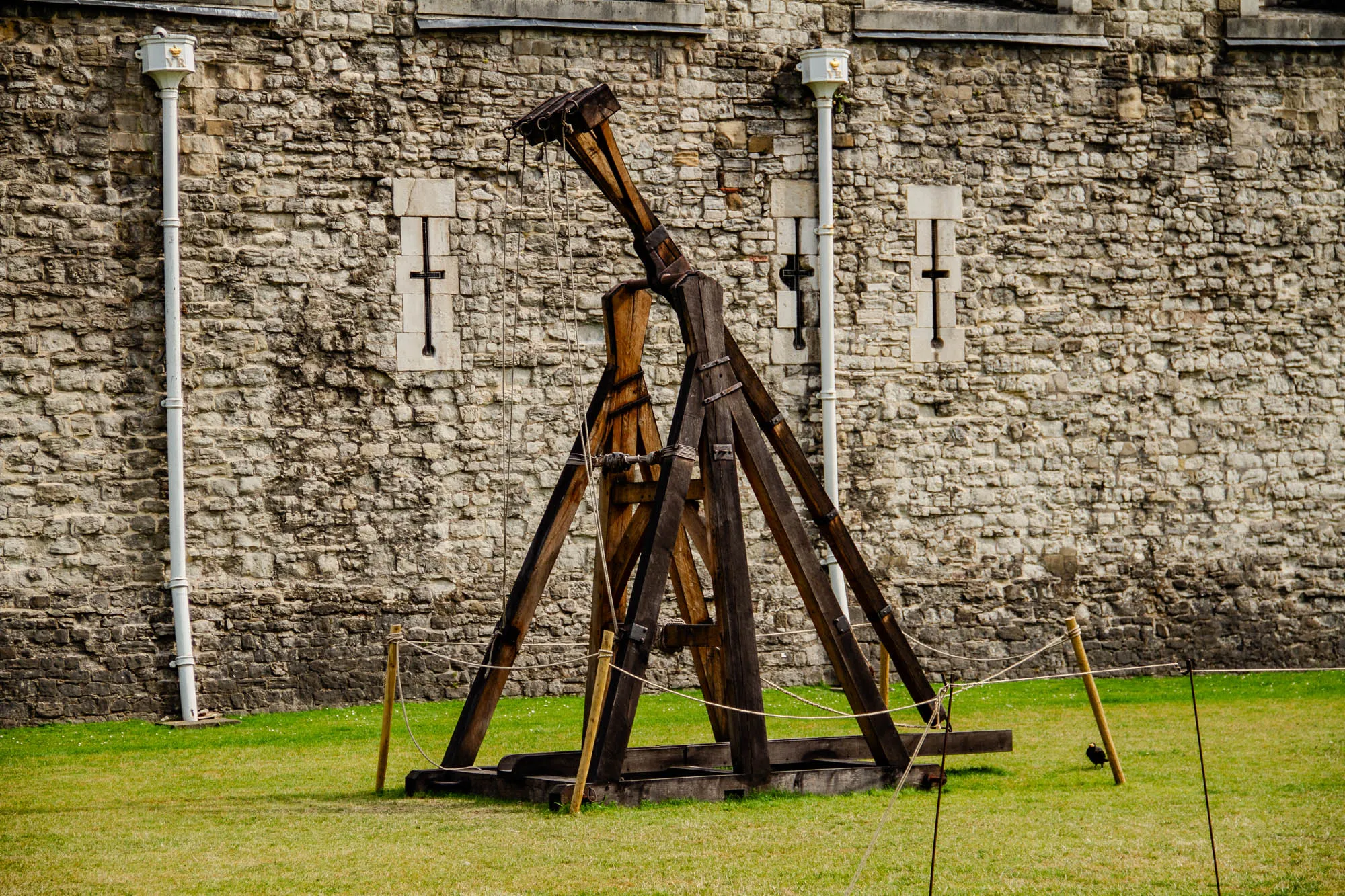 A wooden trebuchet, a siege weapon used to hurl projectiles, stands in front of a stone wall. The trebuchet is made of large, sturdy beams of wood held together with ropes and metal brackets. It has a large arm that swings back and forth, powered by a counterweight.  The wall is made of rough, uneven stones and has two small windows with iron cross shapes. Two white pipes run down the wall, each with a small rectangular structure at the top.