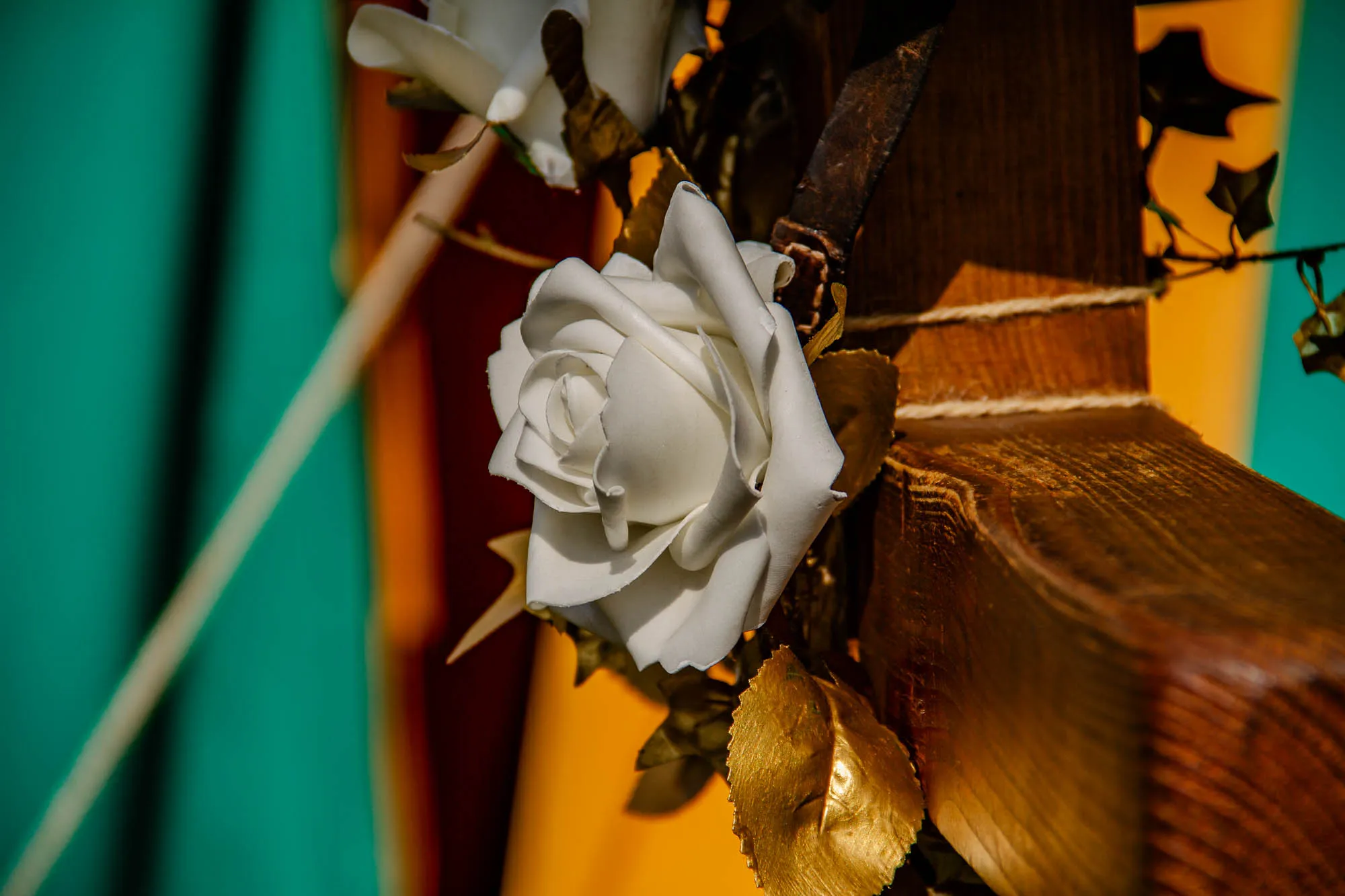 A white silk rose with a wooden stem and gold leaves is attached to a large, weathered piece of wood. The wood is very dark brown, almost black in places, and has a rough texture. There is a piece of twine tying the rose to the wood, and a  piece of brown leather is visible behind the flower.  The wood appears to be part of a larger structure, but only the bottom section is visible in the image. To the right of the rose, there is a blurred green object, and behind the rose is a blurry orange and red object.  The background is very blurry.  The rose is in sharp focus.  The lighting is bright and the image appears to be taken outdoors.