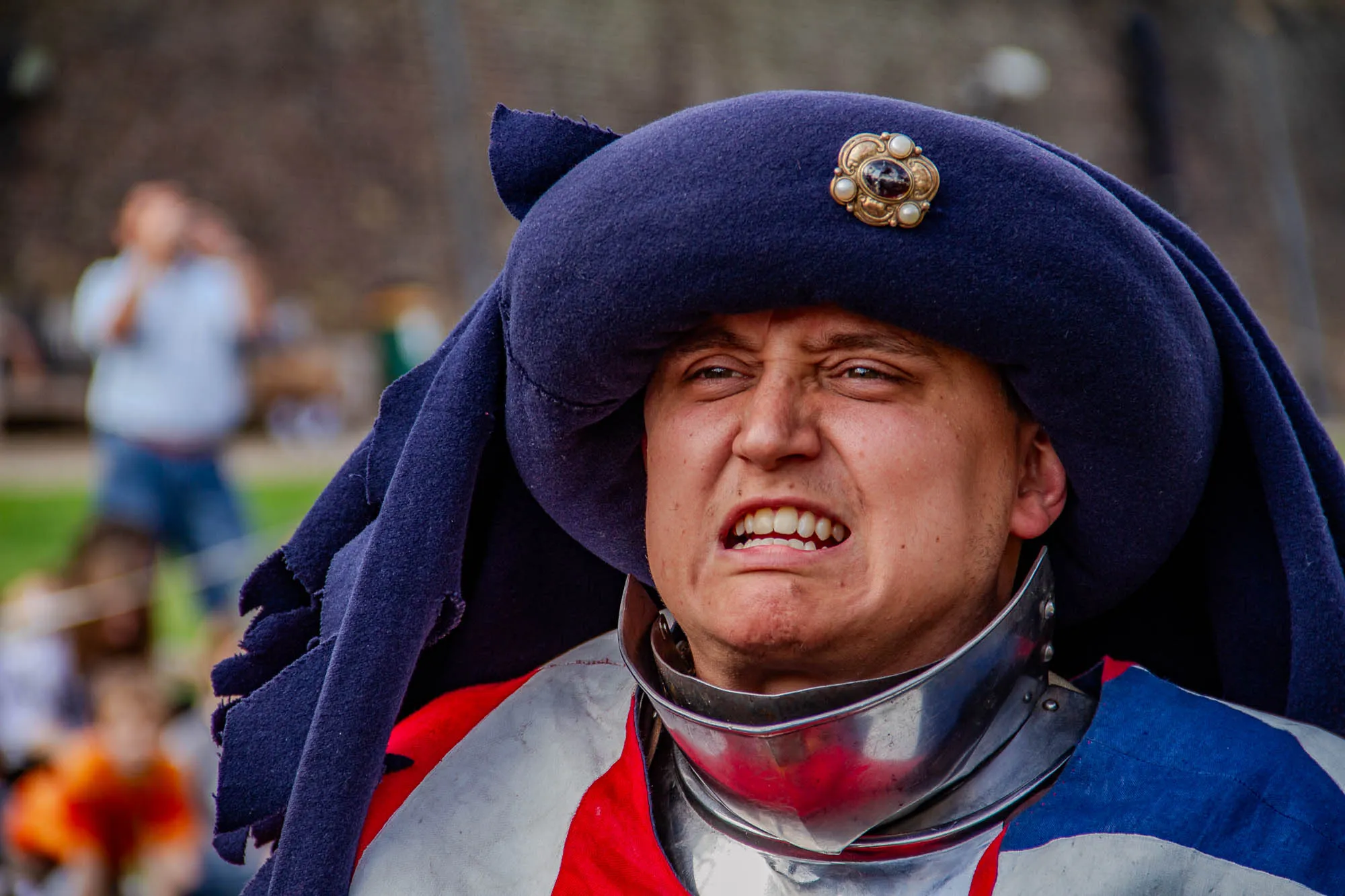 The image shows a man wearing a blue chaperon (medieval hat) with a jewel-encrusted brooch. He's also wearing a suit of armor, with the collar and lower portion of the helmet visible underneath a red and white tunic with a blue stripe on the shoulder. He's looking directly at the camera with a grimace on his face and his teeth are slightly bared, intimidating his opponent. Behind him, in the blurred background, are a few people.  There's also a bit of green grass and a stone wall in the background.