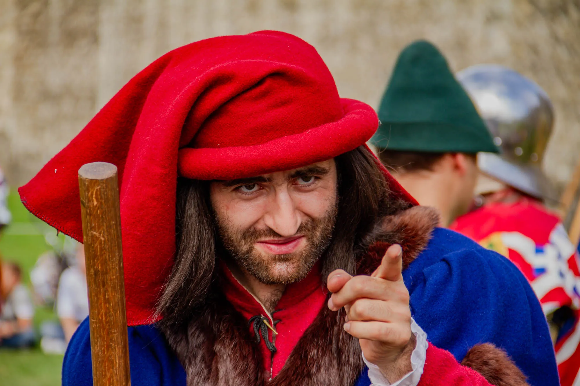 A man with long brown hair and a beard is wearing a red hat, a blue jacket with brown fur trim, and a red shirt. The hat is pulled down over his forehead. He is pointing his right index finger towards the camera. He is holding a wooden staff in his left hand. Another man with a green hat and a red jacket with white accents is standing behind him. The man with the green hat is facing to the left. There is a stone wall in the background.