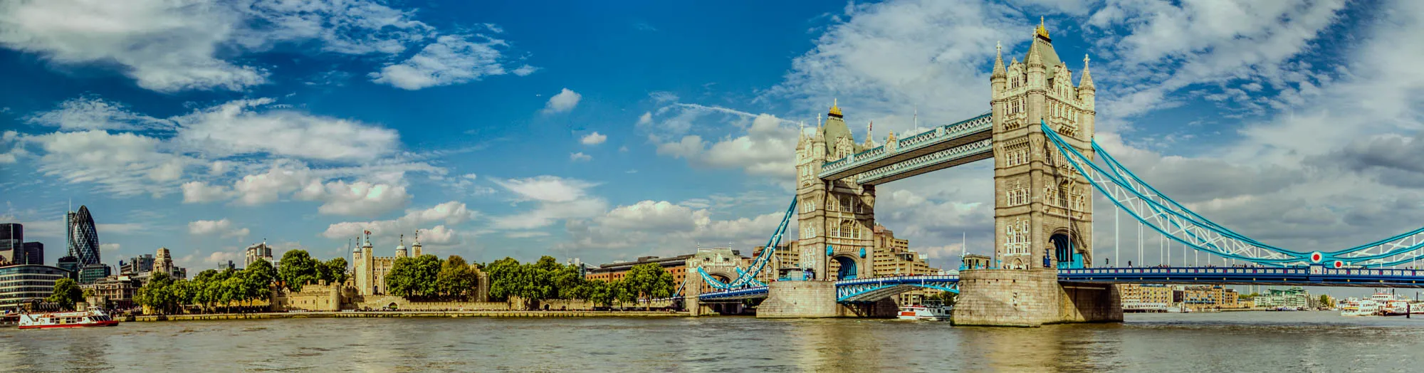 The image is a panoramic view of the Tower Bridge in London, England. The bridge spans a wide river with calm water reflecting the blue sky and white clouds. The bridge is made of two towers, each with a high pointed spire, connected by a walkway. The walkway is made of metal and is decorated with blue and white paint. There are cars and pedestrians walking on the bridge. In the background, you can see the London skyline, including modern skyscrapers and historic buildings and the Tower of London. There is also a boat in the foreground on the river. The overall feel of the image is bright and sunny, with a clear blue sky and fluffy white clouds.