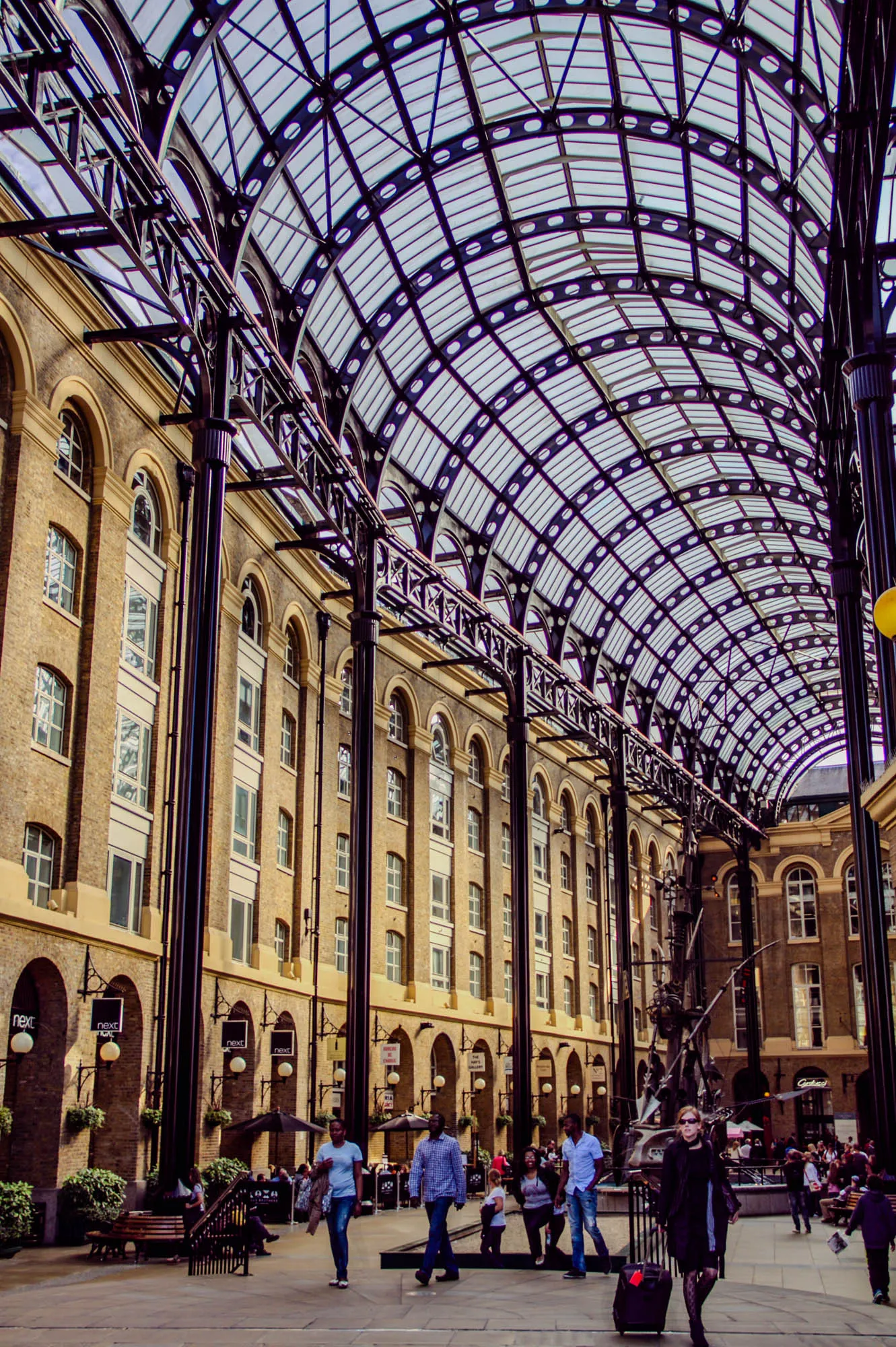 The image shows a wide shot of a large, open, airy space with a metal and glass arched roof. The roof is made of a grid of thick metal beams with glass panels in between. The beams are painted black, and the glass panels are translucent, allowing light to filter through. The space is enclosed by a brick building with tall arched windows. There are several pillars on either side of the space, all of which are painted black. The pillars appear to be the same material as the beams and are likely supporting the roof. There are various people walking through the space, and there are a few small businesses with signs that read "Next". The business are located at the base of the building. Behind the building is a large metal sculpture. There is a walkway in front of the building and more people walking on the walkway.  The photo is taken from a slight angle, looking upwards.  It's a sunny day and the lighting is bright, but the photo seems to have a slightly desaturated color palette.  The focus of the image is the metal and glass roof. 