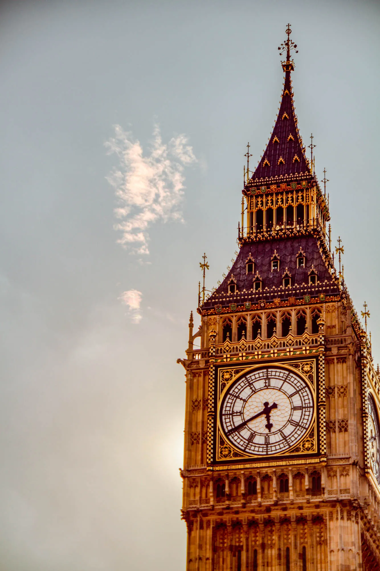 The image shows the Elizabeth Tower, also known as Big Ben, in London.  The tower is a tall, stone structure with a clock face on each side. The image is taken from a low angle, looking up at the tower. The top of the tower is a spire with a flagpole. The clock face is visible in the middle of the tower. There are some white, puffy clouds in the sky behind the tower.  The sky is light blue.  The sun is shining brightly, and it is casting a warm glow on the tower. The tower is a popular tourist destination and is one of the most recognizable landmarks in London.