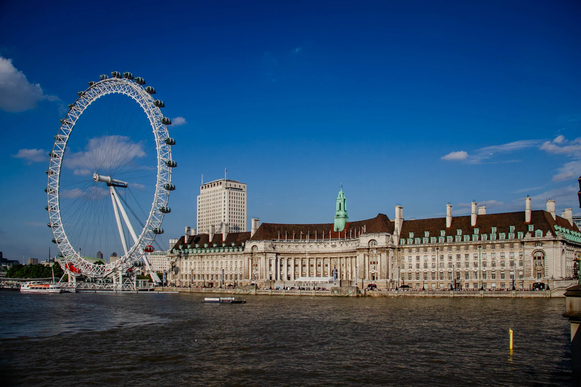 The image shows the London Eye, a large Ferris wheel, standing on the left side of the image. The Eye is white and has a large black frame, and it's located next to the River Thames; it is calm and reflecting the blue sky.  There are people walking around at the base of the wheel, and a boat is docked next to it.  On the right side of the image, you can see a tall white building behind a large, long building with brown roof and many windows. This is the County Hall building. There is a small tower with a green roof on the building. The sky is a clear blue with a few white clouds.  The overall image is bright and sunny. 
