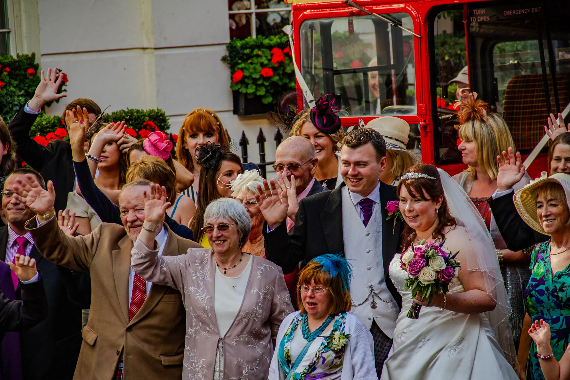 The image shows a wedding party, likely in a city setting. There is a large red double-decker bus in the background. There are many people in the foreground, including the bride and groom. The bride is wearing a white dress and a veil and is holding a bouquet of flowers. The groom is wearing a dark suit and a white vest. He is wearing a white flower in his lapel. Some of the guests are waving at the camera. One guest in the back is standing in front of the bus, waving and wearing a white suit. A guest in the front, wearing a tan suit and red tie, is waving and appears to be very happy. There is a woman in the back wearing a blue dress and a woman in front wearing a pink floral jacket. There is a woman in the front in a floral dress with a turquoise headband.