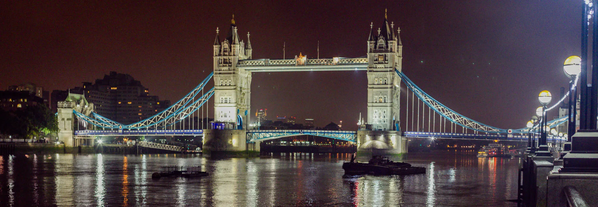 The image shows a nighttime view of the Tower Bridge in London. The bridge is lit up, with its two towers and suspension cables clearly visible. The river Thames is calm, reflecting the lights of the bridge. There is a small boat in the river in the foreground.  In the distance, you can see other buildings and lights. To the right side of the bridge, there are some street lamps illuminating the walkway on the bridge.  