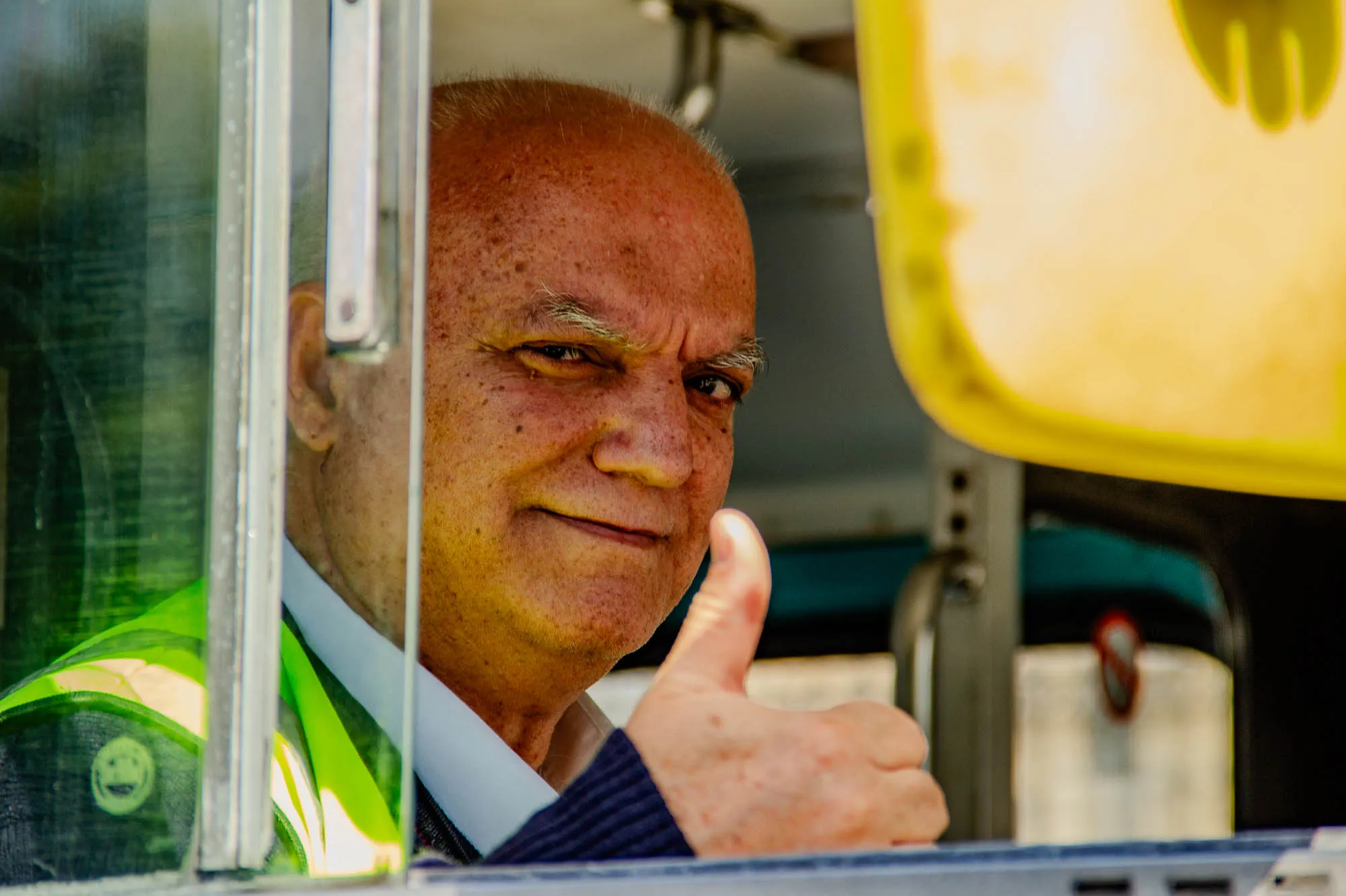 The image is a close-up portrait of a man from the chest up, giving a thumbs up. He is looking directly at the camera. He has a tanned complexion, short, graying hair, and a bit of stubble. He is wearing a blue collared shirt with a white collar, a dark blue sweater, and a bright yellow safety vest with a smiley face logo on it. The man's right arm is visible, with his hand giving a thumbs up to the camera. The image is taken from an interior perspective. The man's left side and arm are mostly obscured by a metal door with a clear window that shows a green and white blur. The top of the frame shows a bright yellow object, possibly a part of the vehicle, that appears to be a yellow safety sign or a piece of equipment.  The interior of the vehicle is also visible.  The background is blurred and out of focus. The overall feel of the image is positive and upbeat.