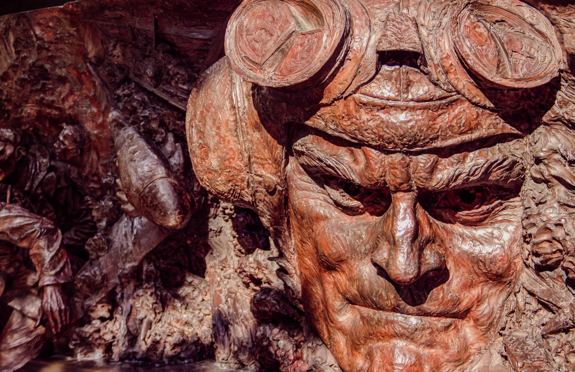 The image is a close-up of a bronze sculpture of a man's face. The man is wearing a pair of goggles with large, round lenses. His face is weathered and wrinkled, and he has a determined look on his face. The sculpture is very detailed, and the artist has captured the man's personality and spirit in the work.  The background is a rocky, textured surface, perhaps representing a battlefield or a scene of war. This sculpture is a powerful reminder of the sacrifices made by those who fought in wars.  The bronze is a reddish-brown color, and the light catches the surface of the sculpture, creating highlights and shadows that give the sculpture a sense of depth and realism. The sculpture is very expressive, and the man's eyes seem to be looking directly at the viewer.