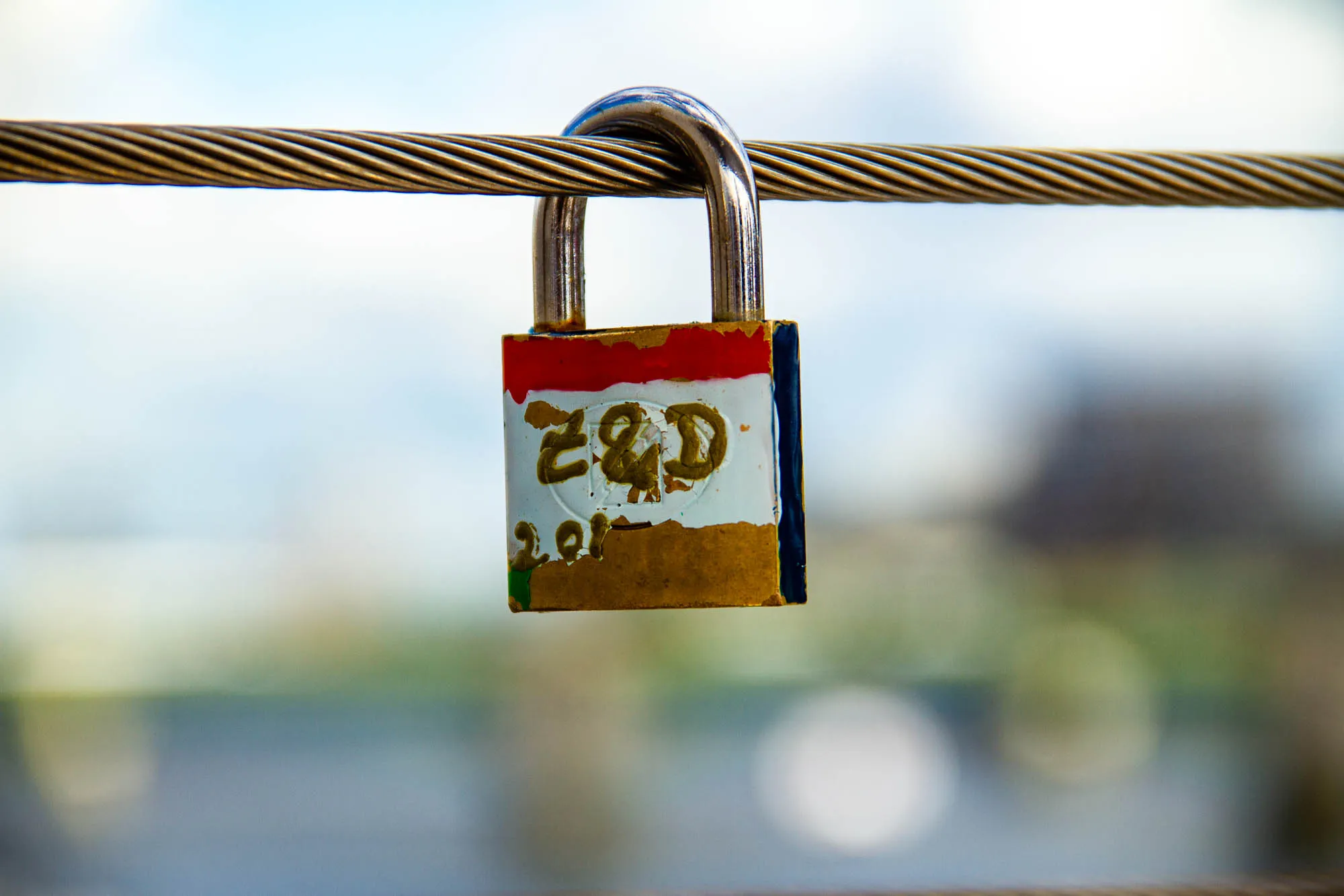 A bronze padlock is hanging from a thick cable. The padlock is closed and is painted red, white, and black. On the front is handwritten in gold paint "Z&D" and "2010", with the "0" mostly cut off. The background is a very blurry light blue, with a dark grey blur on the upper right and a lighter green blur below it.