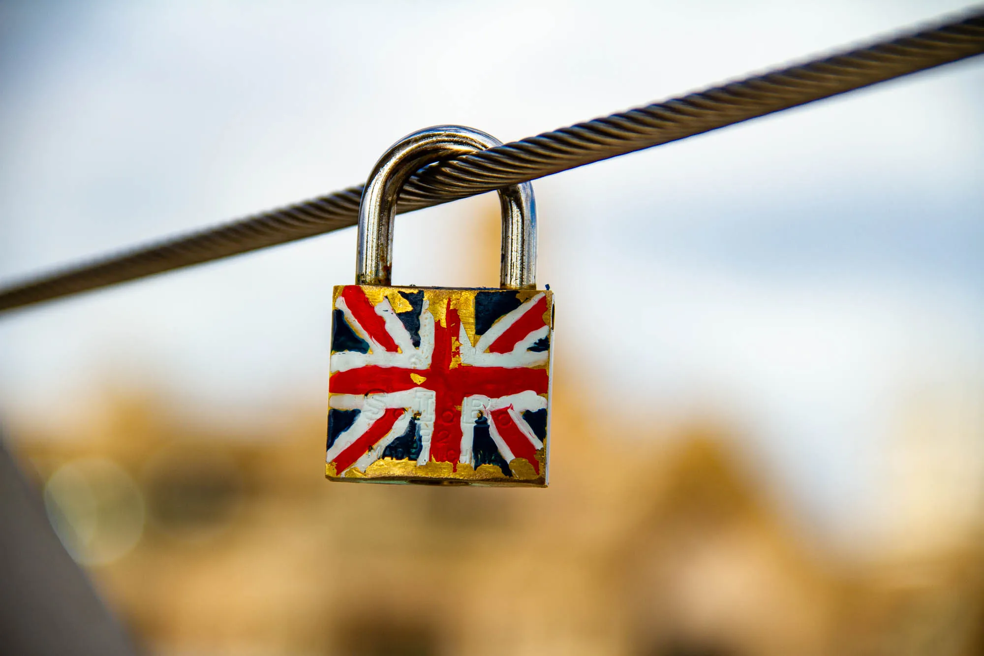 The image shows a padlock hanging from a steel cable. The padlock is painted with the Union Jack, the flag of the United Kingdom. The colors of the flag are red, white, and blue. The background is blurred and out of focus, but it appears to be a city scene with some buildings in the background.  