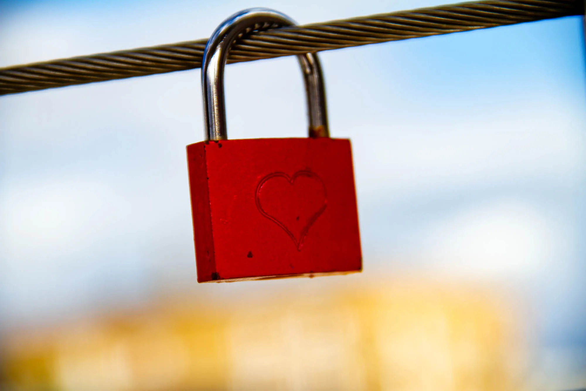 A red padlock with a heart engraved on it is hanging from a thick metal cable. The padlock is in focus and the cable is slightly out of focus. The background is out of focus and appears to be a blue sky with some yellow and orange hues. 