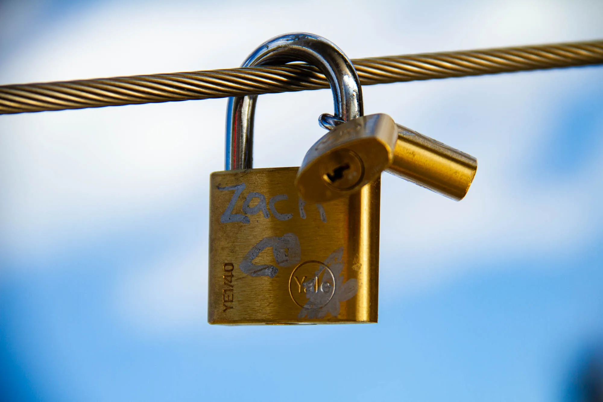 The image shows a close-up of two gold padlocks attached to a thick, braided cable. The padlocks are both closed and are hanging from the cable. The padlock on the left is larger and has the word “Yale” inscribed on it in a circular shape. It also has the word “Zach” and a heart inscribed on it. The padlock on the right is smaller and appears to be older and more worn. The cable is attached to a blurred background that appears to be a bright blue sky. 
