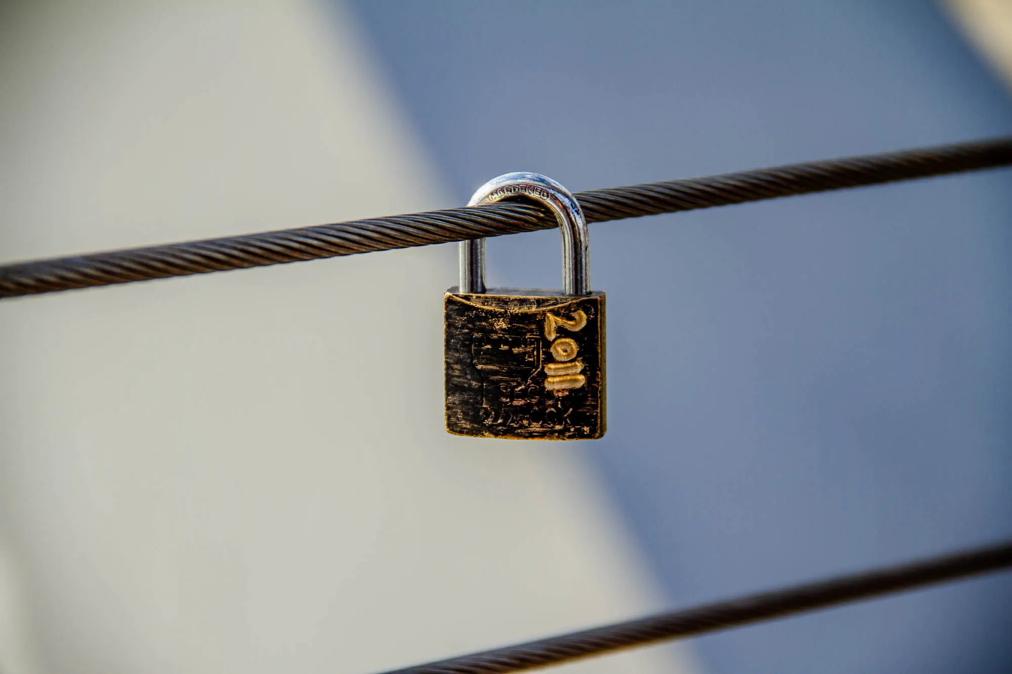 A black padlock with a silver shackle is attached to two parallel metal cables. The padlock has a small engraved design and the year 2011 is visible on its surface.  The background is a blurred light gray and dark blue.