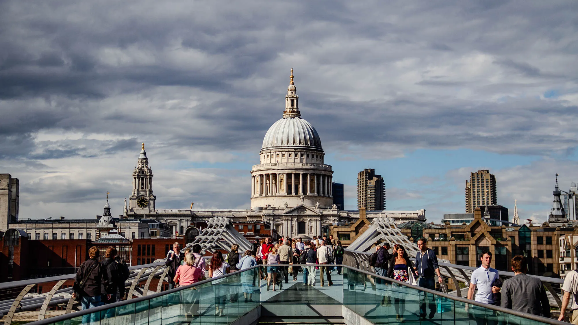 This is a photo of a busy street in London, England.  In the center of the image is St. Paul's Cathedral, a large domed church.  In the foreground there are many people walking on a glass bridge and there are buildings all around the scene.  The sky is cloudy, but the sun is shining. The scene is vibrant and full of life.