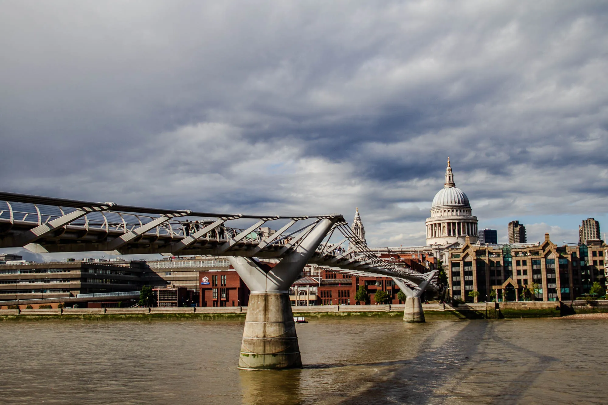 The image shows a view of the Millennium Bridge in London, England. The bridge is a modern pedestrian bridge that spans the River Thames.  It is a suspension bridge with a distinctive curved shape and a steel deck. The bridge is made up of two large piers and a series of connecting beams. In the image, the bridge appears to be crowded with people walking across.  The River Thames is in the foreground and is calm and reflective. In the background, you can see the dome of St. Paul's Cathedral, a prominent landmark in London.  The sky is overcast with grey clouds.  The image captures a moment of urban life in London, showcasing a modern architectural landmark against a backdrop of historic buildings. 
