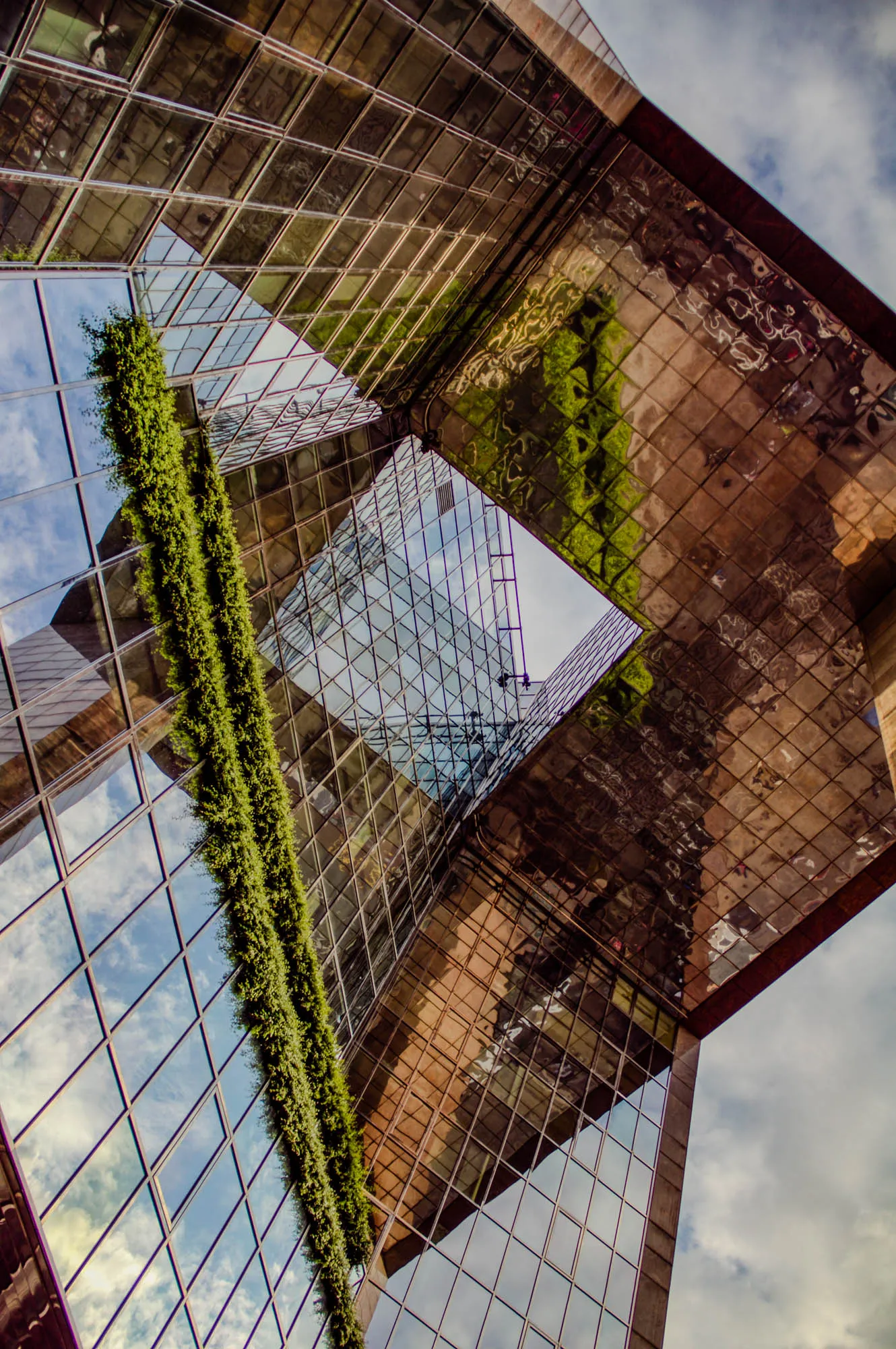 The image shows a low angle view of a modern glass building. The building is made up of a series of large windows with a grid pattern. A vertical strip of green plants hangs down along the side of the building. The windows are reflective, and they reflect the sky and other buildings. A large skylight, shaped like a diamond, allows natural light to pour in from the roof.  The sky is mostly cloudy and white. The overall feeling is one of modernity and sophistication.  
