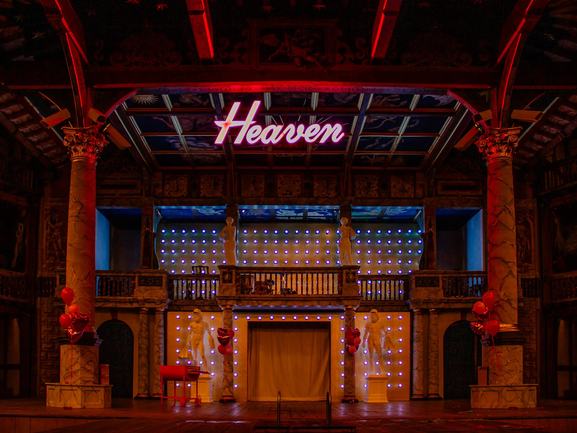 The image shows a stage with a large neon sign reading "Heaven" above a dark, ornate, painted backdrop. The stage is lit with a grid of blue lights and has several large white pillars running along the sides, with a marble railing and a dark archway in the back.  A few red balloons are attached to the bottom of the left pillar.  There are two statues of nude men on pedestals in front of the back wall.  The stage is made of wood and appears to be set up for a performance.