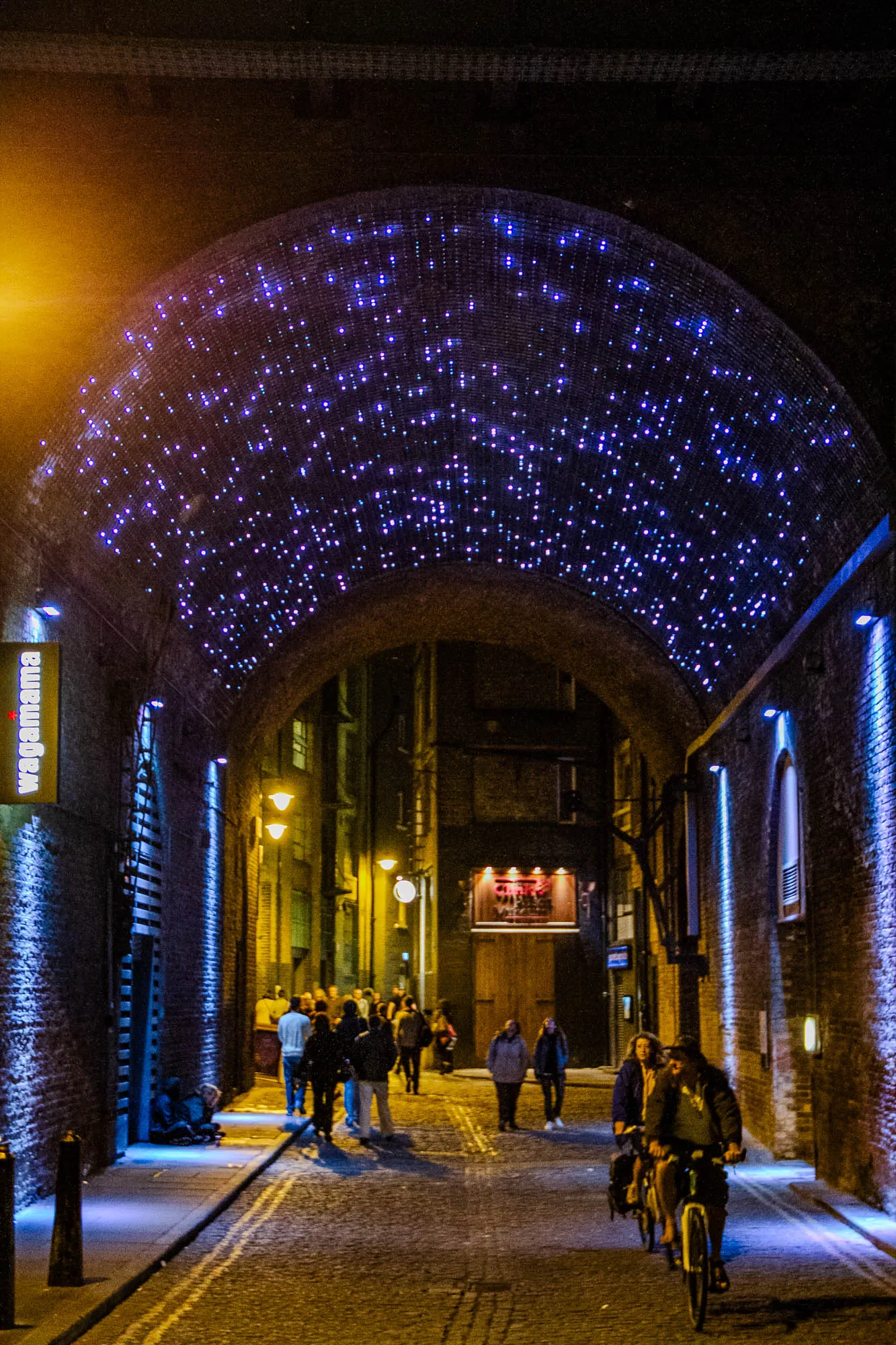 This is a night time image of a narrow underpass, with brick walls on either side. The street is lit by string lights that have been strung across an archway over the street, resembling a starry night sky. In the foreground, there is a couple riding a bicycle toward the viewer. Behind the couple is a group of people walking along the street. Further down the street, a sign can be seen for a restaurant called "Wagamama" and another sign can be seen for the "Clink Prison Museum". The street is paved and the brick walls are illuminated by blue lights. 
