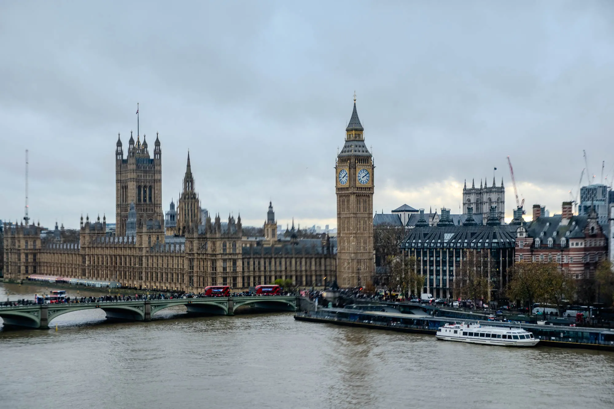 The image shows a view of the Houses of Parliament and Big Ben in London, England. In the foreground, a bridge crosses the River Thames, with a white boat docked on the right side of the river. Several people are walking across the bridge, and there are two red double-decker buses on the bridge. The image is taken from a high vantage point, looking down at the river and the city beyond. The sky is cloudy, and the buildings in the background are mostly out of focus. Big Ben, the iconic clock tower, stands tall in middle, its face visible. To the left of the tower is the Houses of Parliament, a large complex of buildings that have served as the home of the UK Parliament since 1800.