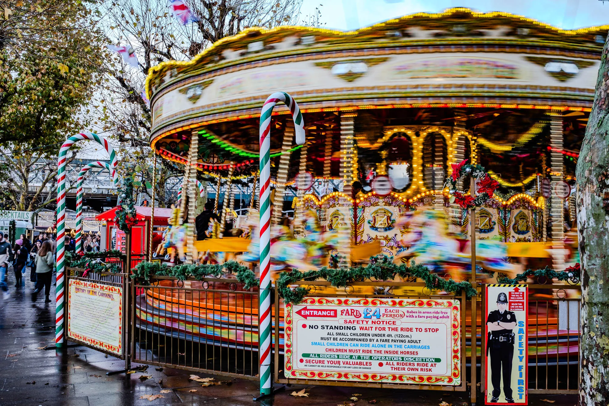 The image depicts a brightly colored carousel with lights and a sign that says "Fare £4". The carousel is in motion, indicated by the blurry figures of people riding and the blurred lights. There are several large candy canes striped in red and white at the front of the carousel, and it is surrounded by a black metal fence. The carousel is decorated with festive greenery and red poinsettia flowers. Next to the carousel is a sign with instructions for riding the carousel, with text reading: "Safety notice, no standing, wait for the ride to stop, all small children under 4ft must be accompanied by a fare paying adult," and more. Next to this sign is a sign depicting a police officer that says, "Safety first, you must be this tall to ride a horse, unaccompanied below this line can ride in a carriage." To the left of the carousel, there is a tree, and beyond the tree is a large, festive, red building, possibly a storefront, with text that reads, "cocktails."  The ground in the foreground is wet. There are other people in the background, but their appearance is blurred.  