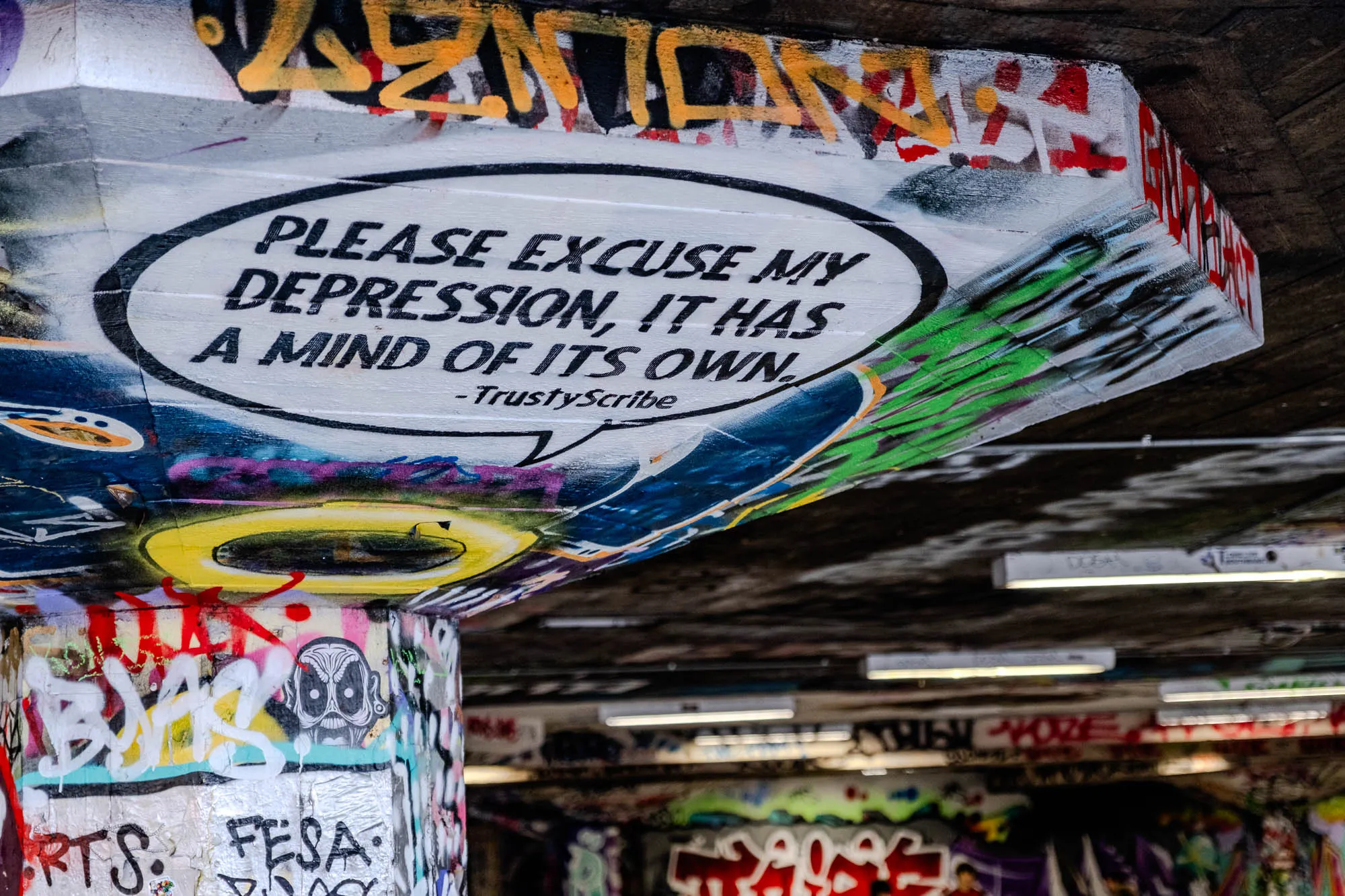 The image is of a concrete pillar covered in graffiti. The top of the pillar is painted white with a black speech bubble on it.  The speech bubble says, "Please excuse my depression, it has a mind of its own." The caption underneath the speech bubble says, "-TrustyScribe."  The rest of the pillar is covered in colorful, random graffiti, including what appears to be a stylized face in the center. There are several fluorescent lights in the ceiling above. 
