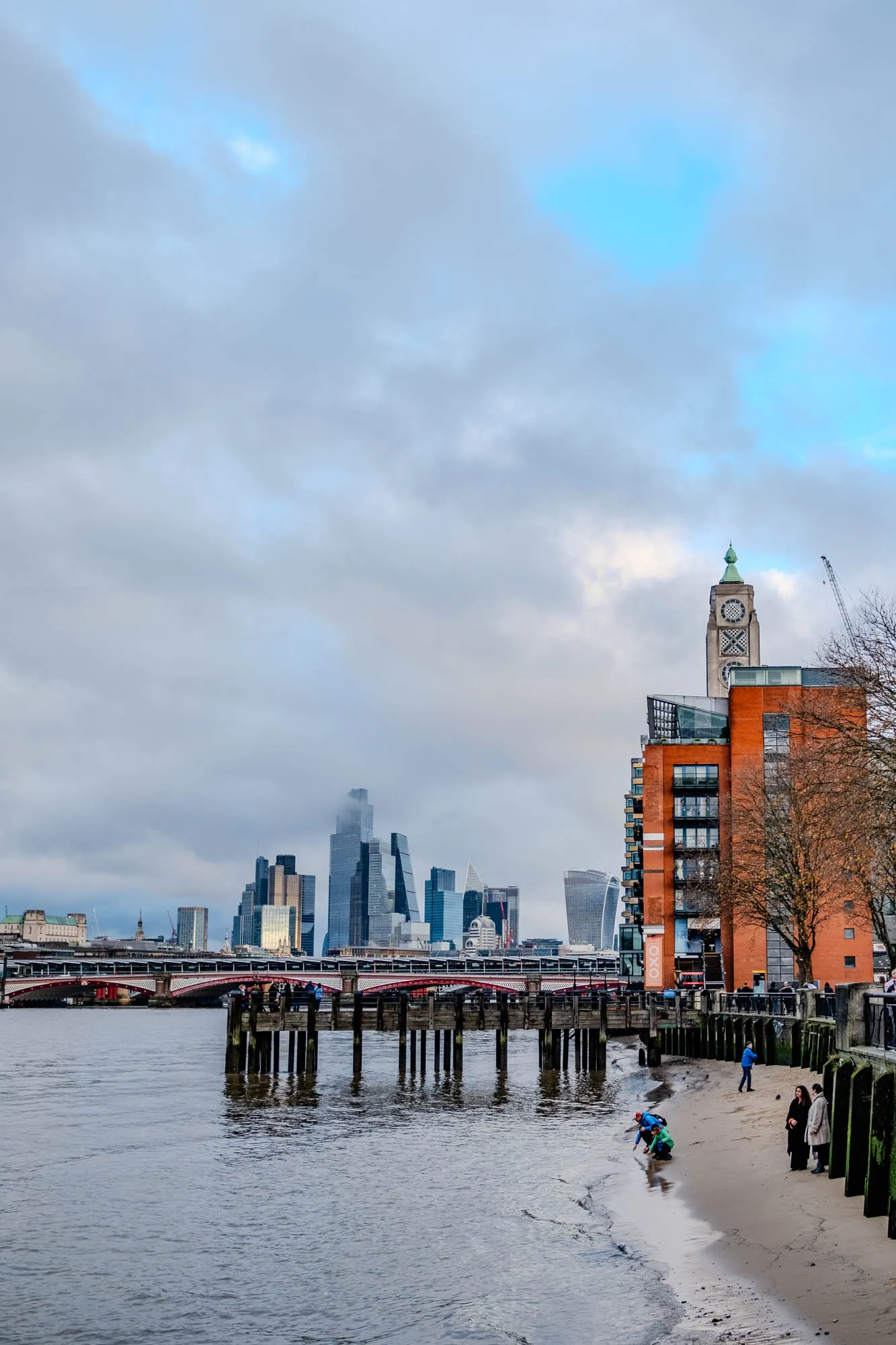 The image shows a view of the River Thames in London. In the foreground, there is a sandy beach with a group of people walking along the shoreline.  A pier juts out into the water, and there are some wooden posts supporting the structure.  A large building with a clock tower and a red brick facade sits on the right edge of the image.  In the distance, there is a city skyline, including several tall skyscrapers.  The sky is overcast, with patches of blue peeking through the clouds. The image is taken from a low angle, looking out across the water.