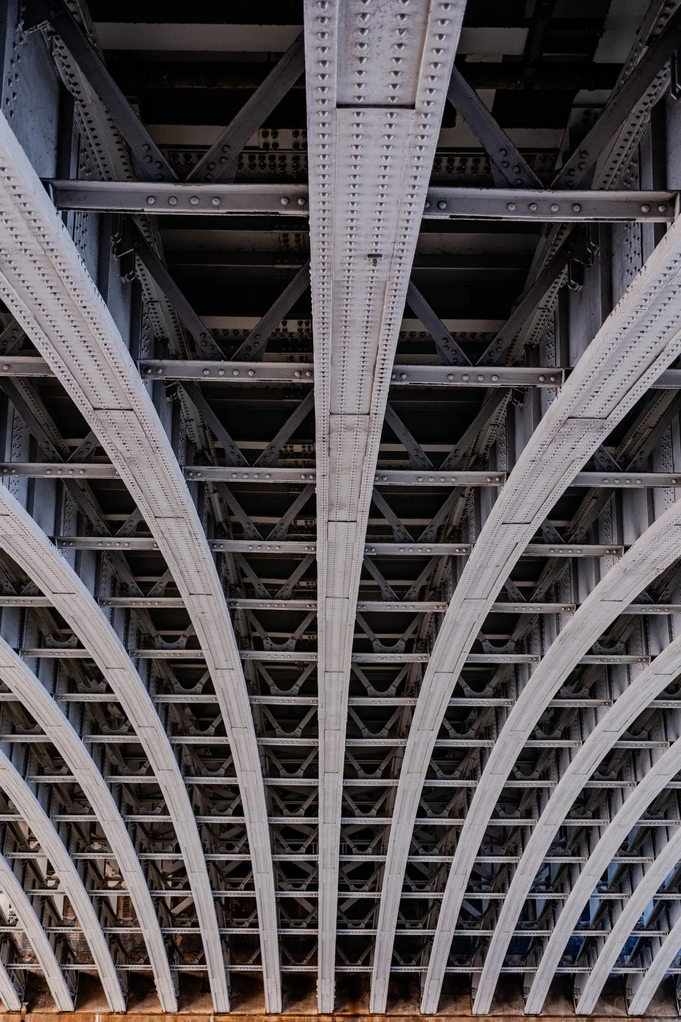 The image shows the underside of a bridge, with many criss-crossing steel beams creating a grid-like pattern. The beams are all painted in a light grey color and have small round rivets along the edges. The beams are arranged in a repeating pattern of arches, creating a sense of depth and perspective. The top of the image is obscured by the beams, and the bottom of the image shows a hint of the concrete supports below. The image is well-lit and the details of the steel structure are clear.