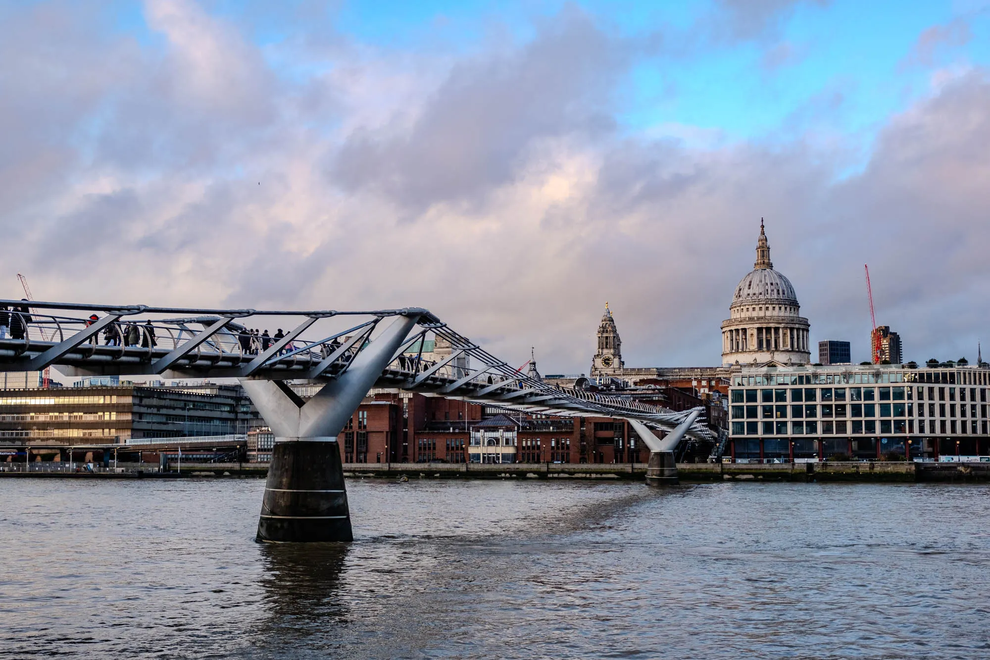 The image shows a view of the Millennium Bridge in London, England. The bridge is a modern pedestrian bridge that crosses the River Thames. The bridge is made of steel and has a distinctive curved shape. There are people walking on the bridge. In the background, the iconic St. Paul's Cathedral is visible. The sky is a cloudy blue with some white clouds. The river is calm and reflective, with the water reflecting the sky and the buildings.